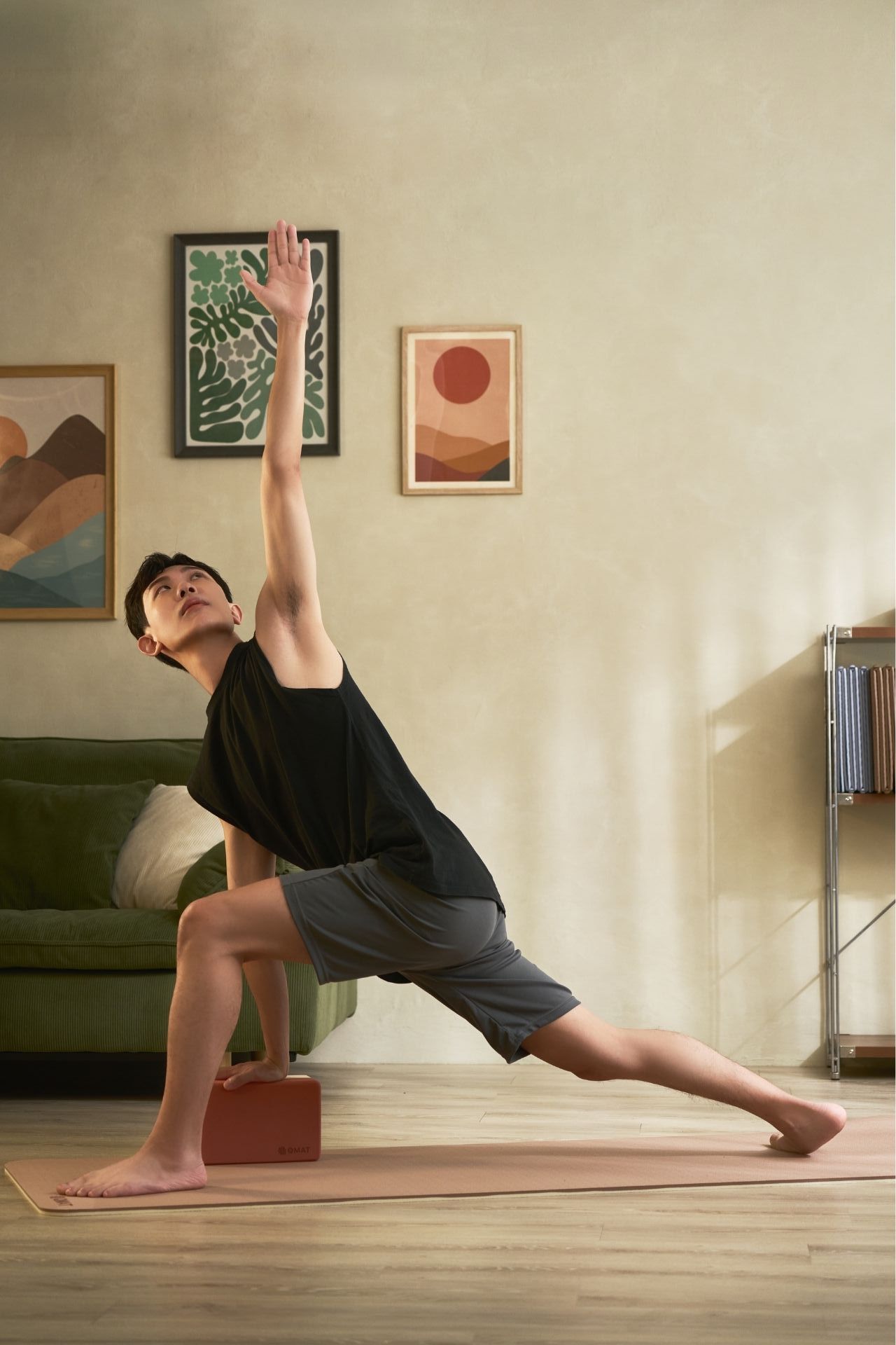 Man practicing yoga side angle pose on mat with block, wearing black tank top and shorts indoors.