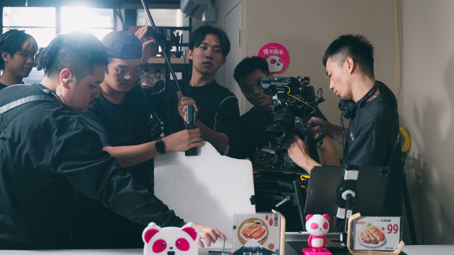 Film crew operating a professional camera on a busy set with colorful props and merchandise displayed on a counter.