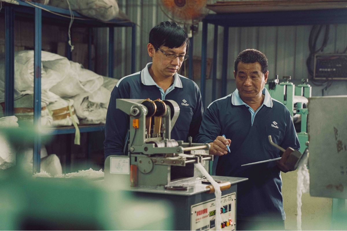 Two workers in blue uniforms examine equipment at an industrial workstation with machinery and tools.