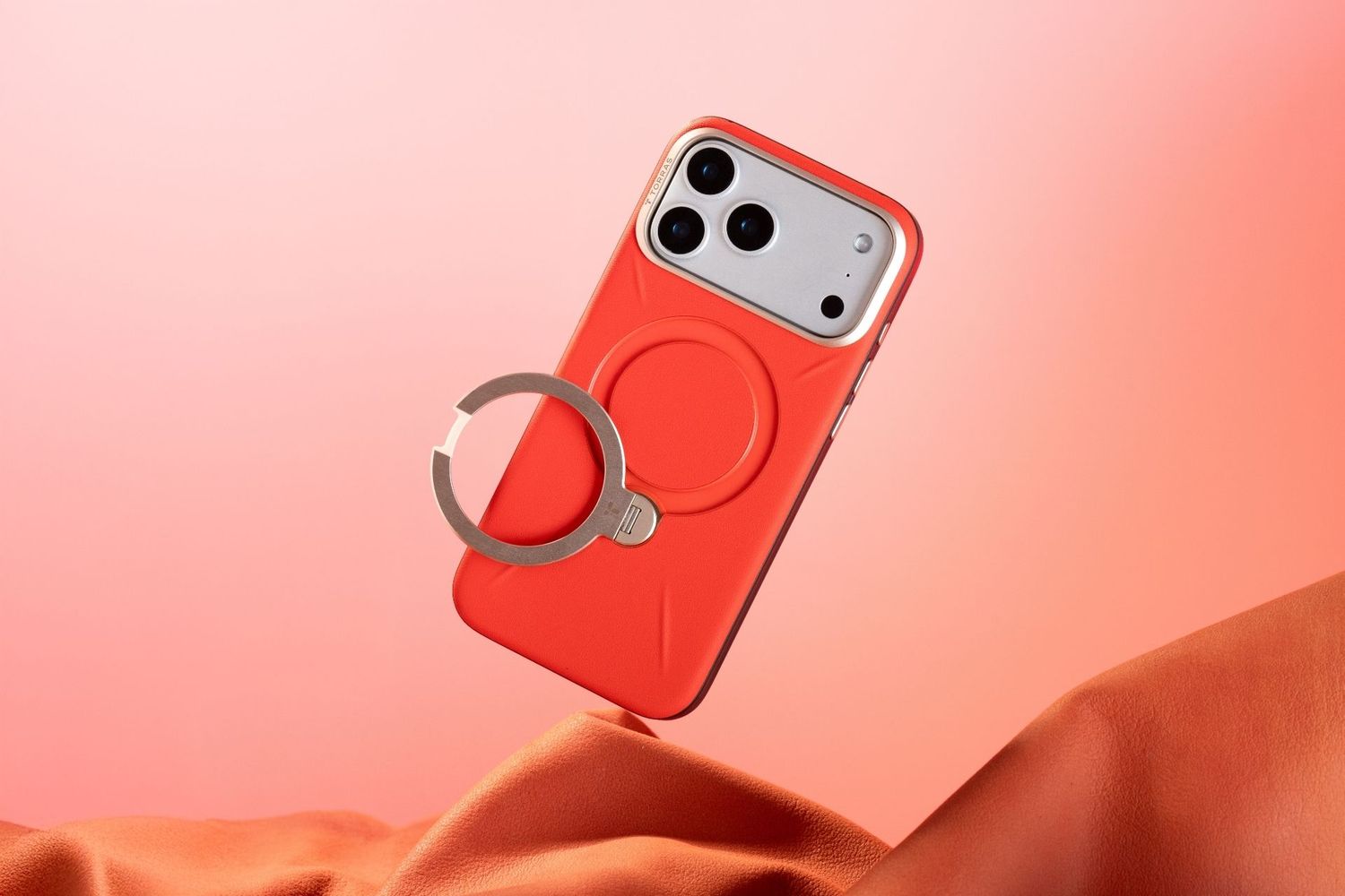 Red MagSafe iPhone case with metal ring stand floating against a coral pink background with silk fabric.