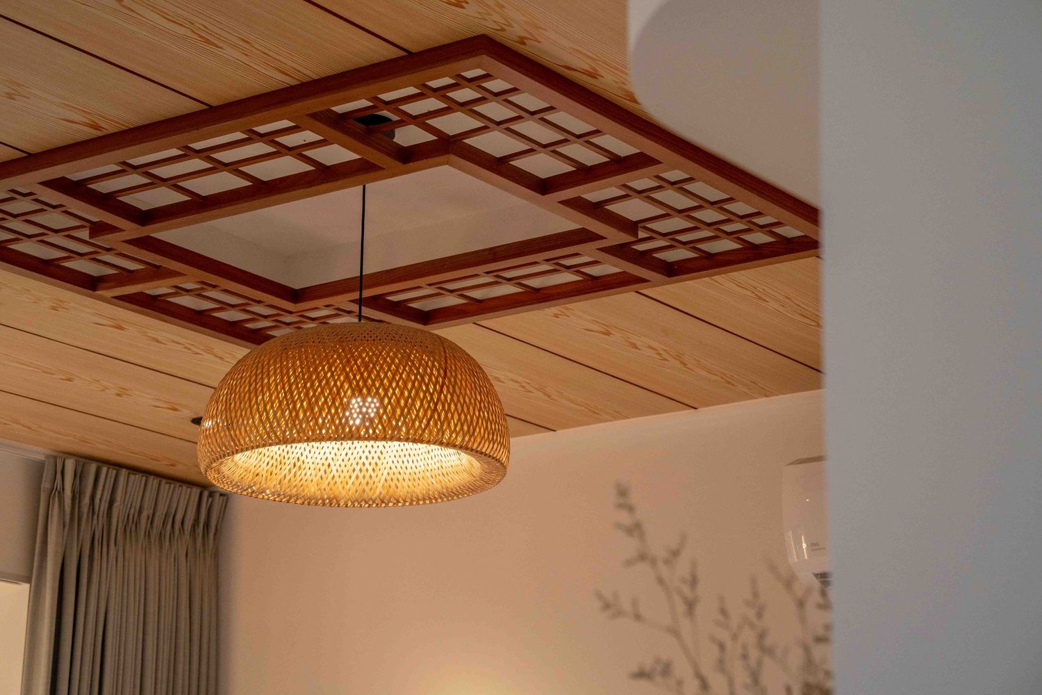 A rattan woven pendant lamp hangs from a wooden ceiling with geometric grid pattern and white curtains visible.