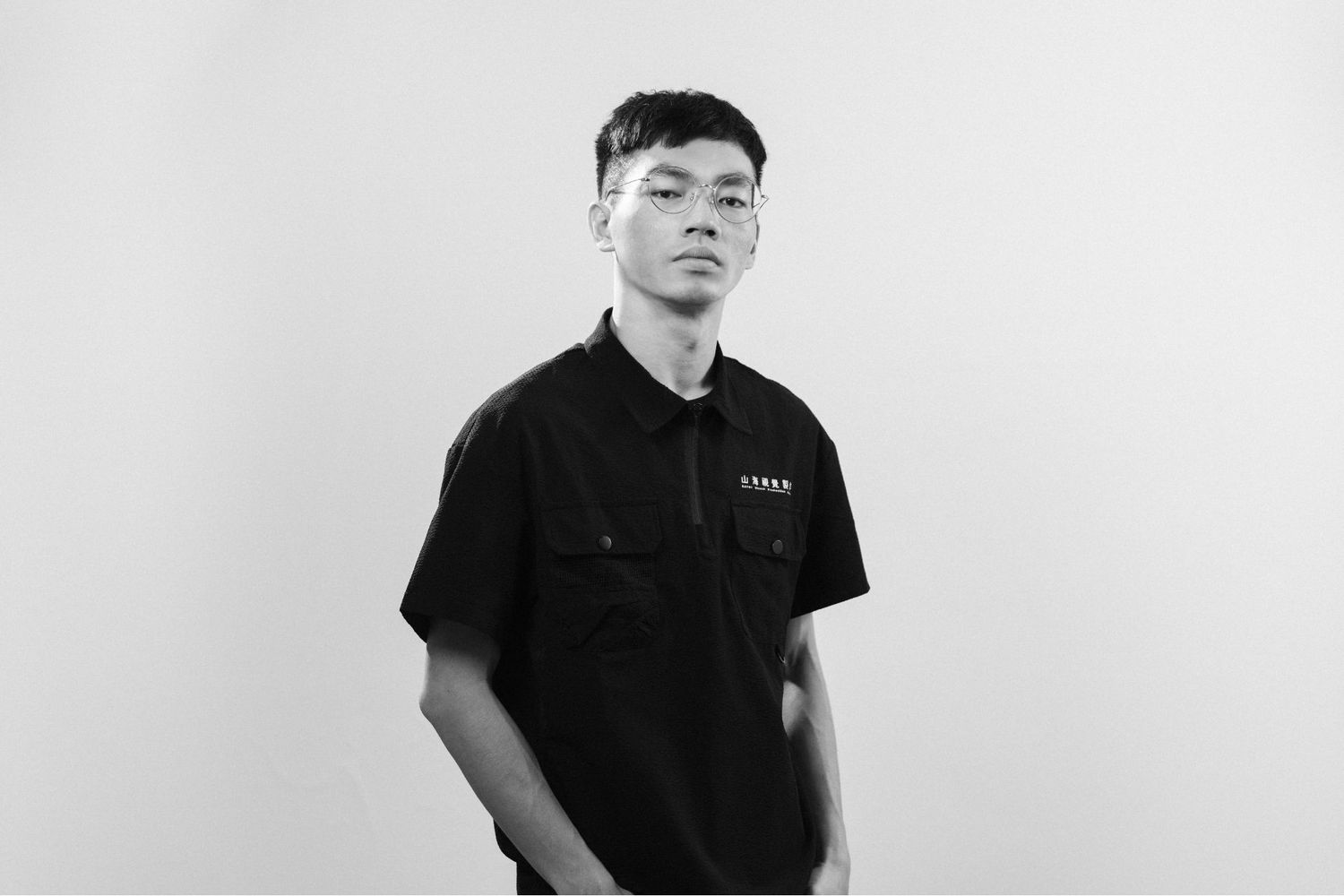 A person in a black polo shirt poses against a plain white background in a minimalist black and white portrait.