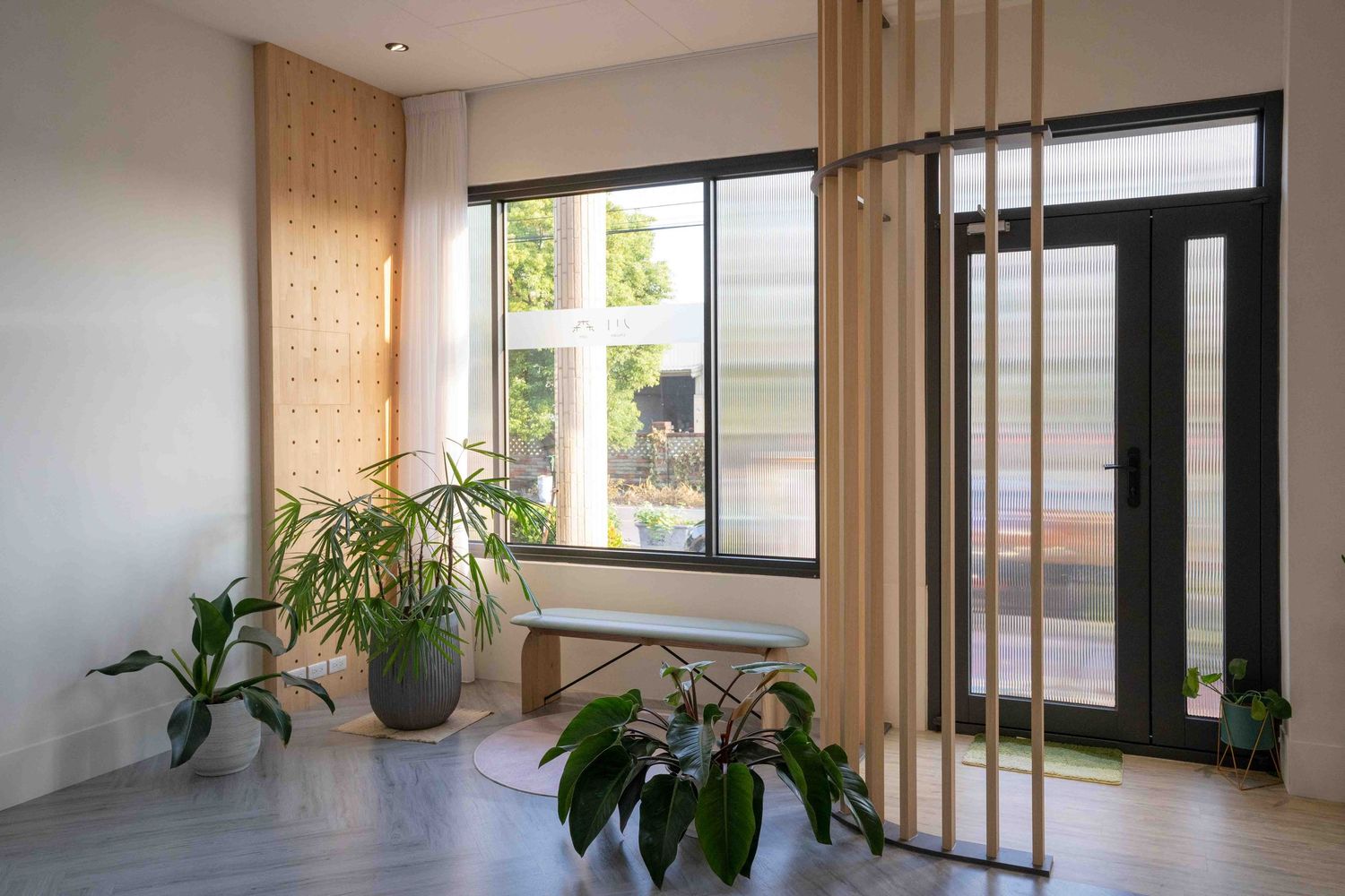 Modern interior space with large windows, wood slat divider, and potted plants beside a glass entrance door.