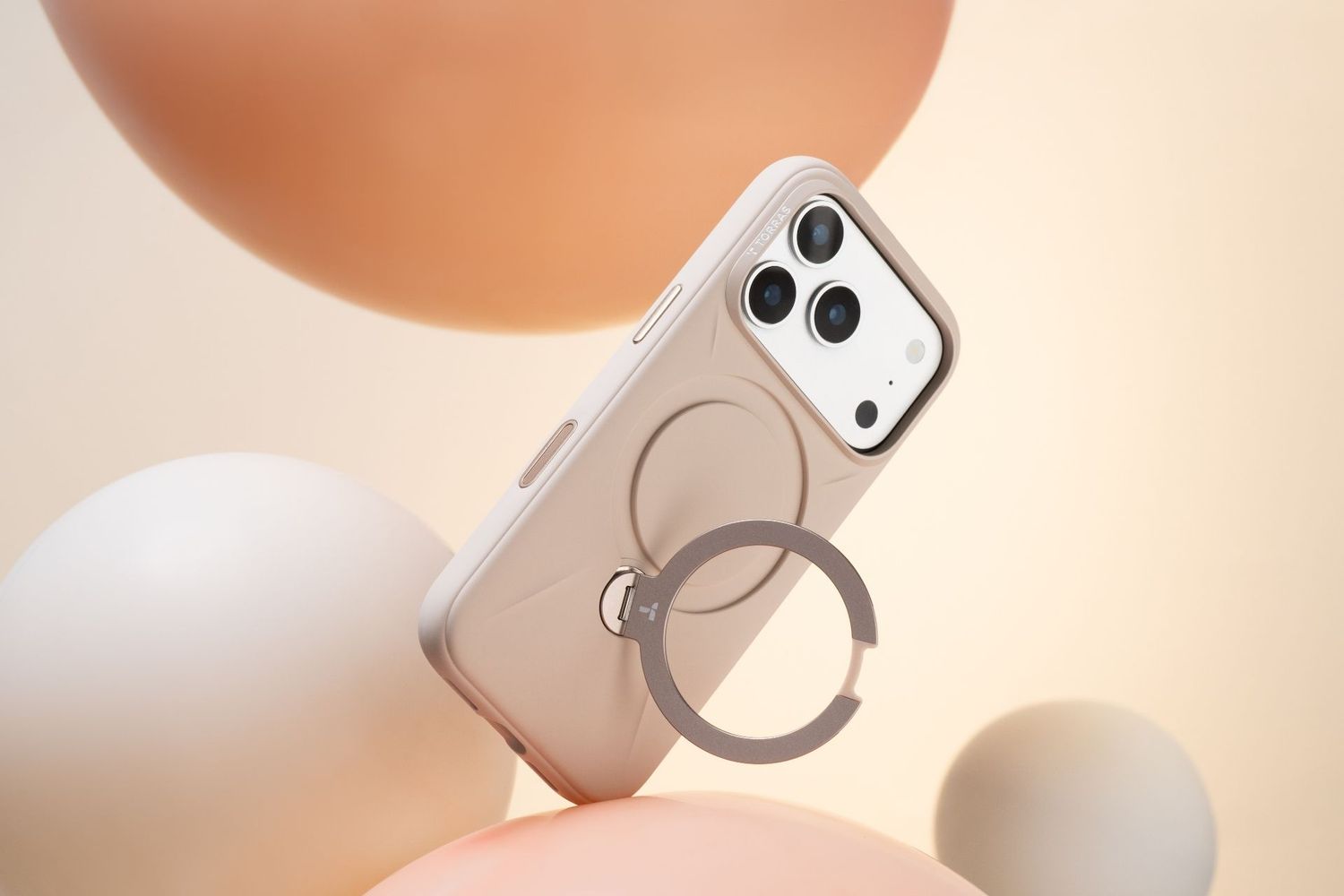 Beige iPhone 15 Pro MagSafe case with ring holder floating among soft pastel spheres on cream background.