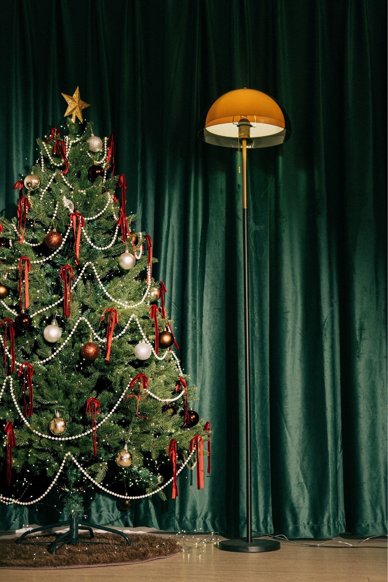 Decorated Christmas tree with gold star, red ribbons, and baubles beside a retro floor lamp against green velvet curtains.
