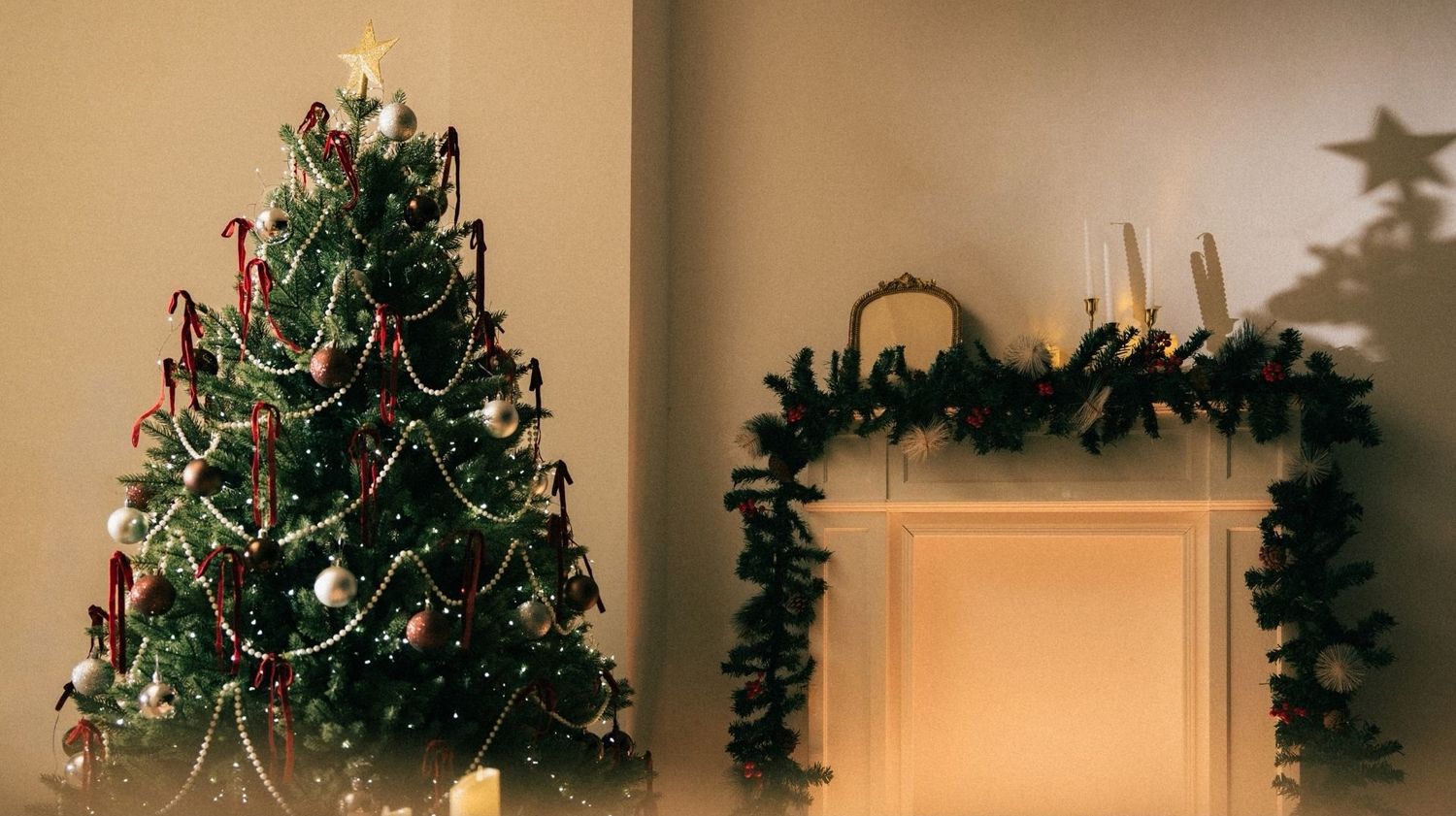 A decorated Christmas tree with red ornaments stands beside a garland-adorned fireplace mantel in a cozy room.