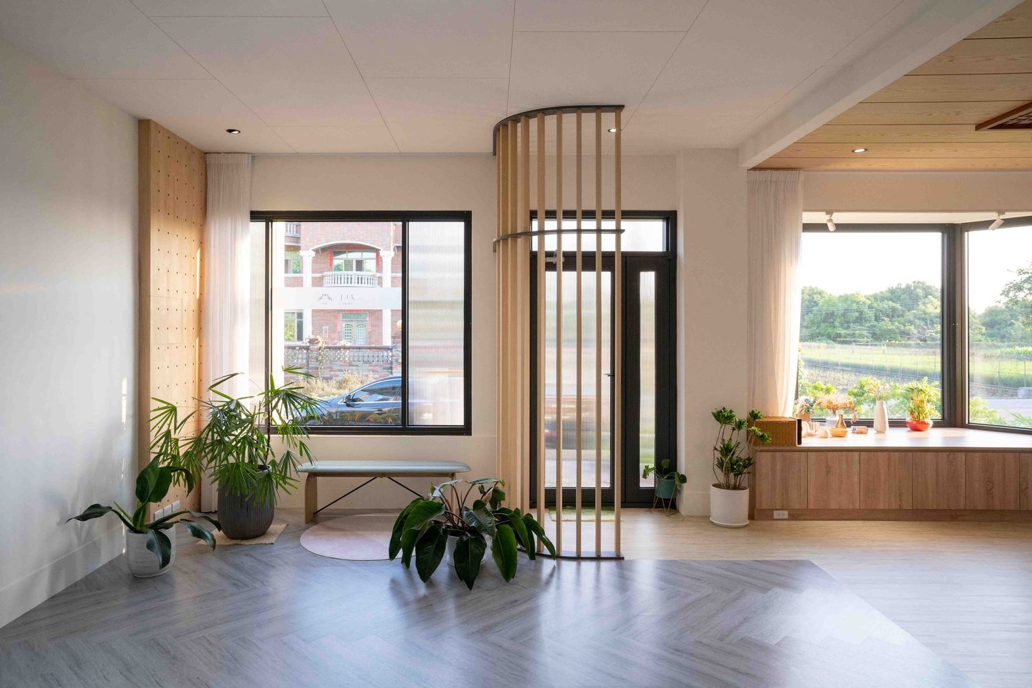 Modern interior space with herringbone flooring, curved metal divider, and potted plants by large windows with natural light.