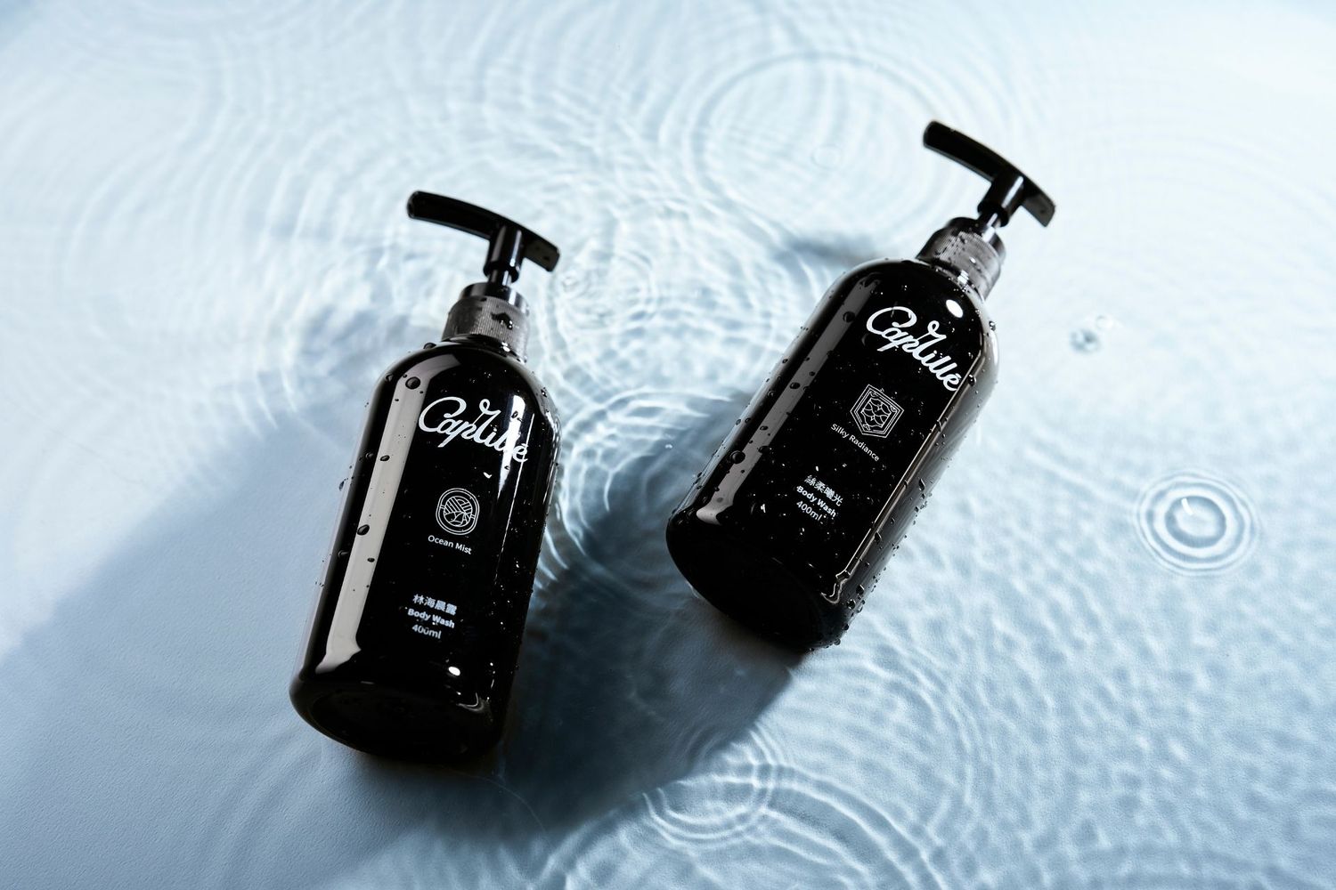 Black glass perfume bottle with silver cap photographed in water with ripple effects and reflections.