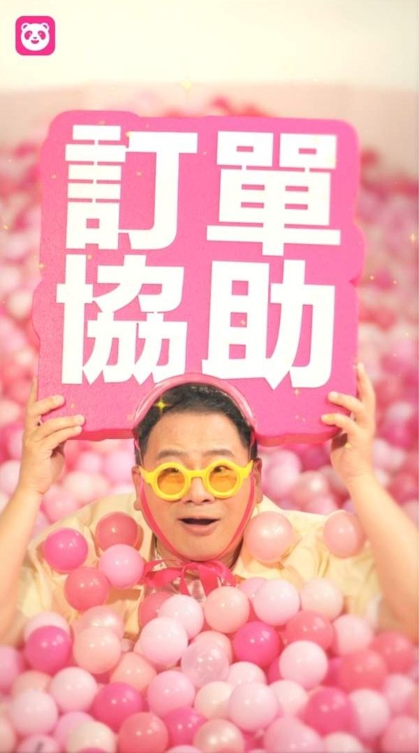 Man in yellow glasses holds pink sign reading order assistance, surrounded by pink balls in a fun foodpanda promotional image.