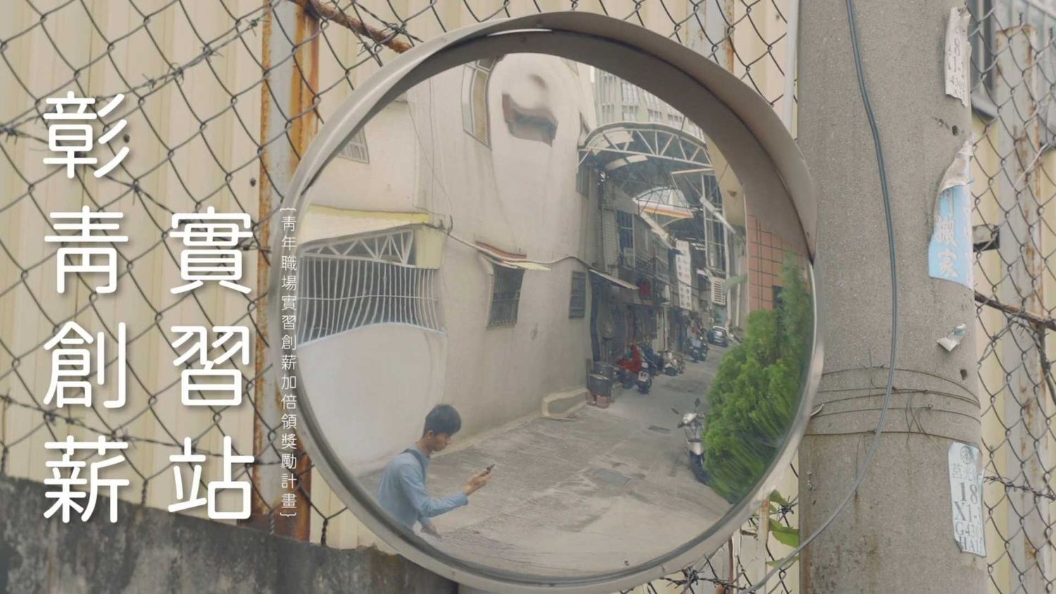 A circular mirror on a wall reflects a narrow alleyway with buildings and utility poles in an Asian neighborhood.