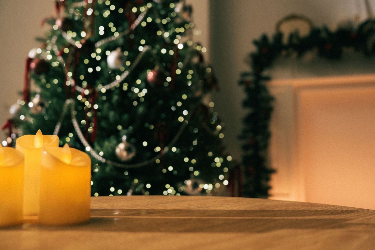 Yellow candles on wooden table with blurred Christmas tree and fireplace with warm bokeh lights.