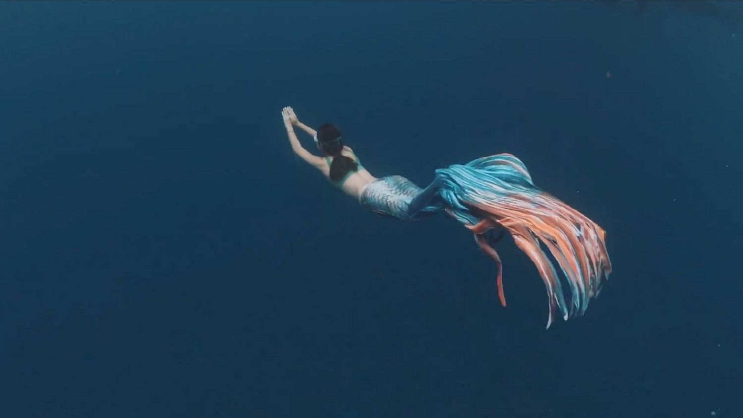 A person in flowing turquoise and coral fabric dives gracefully through deep blue water.
