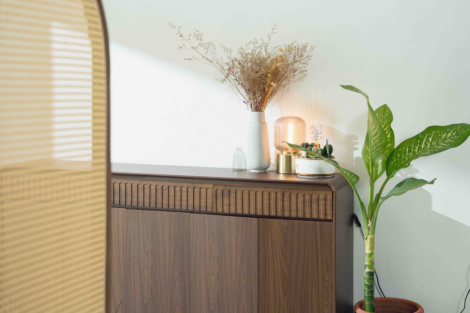 A modern wooden cabinet decorated with a potted plant, dried flowers in a vase, and stylish home decor against a mint wall.