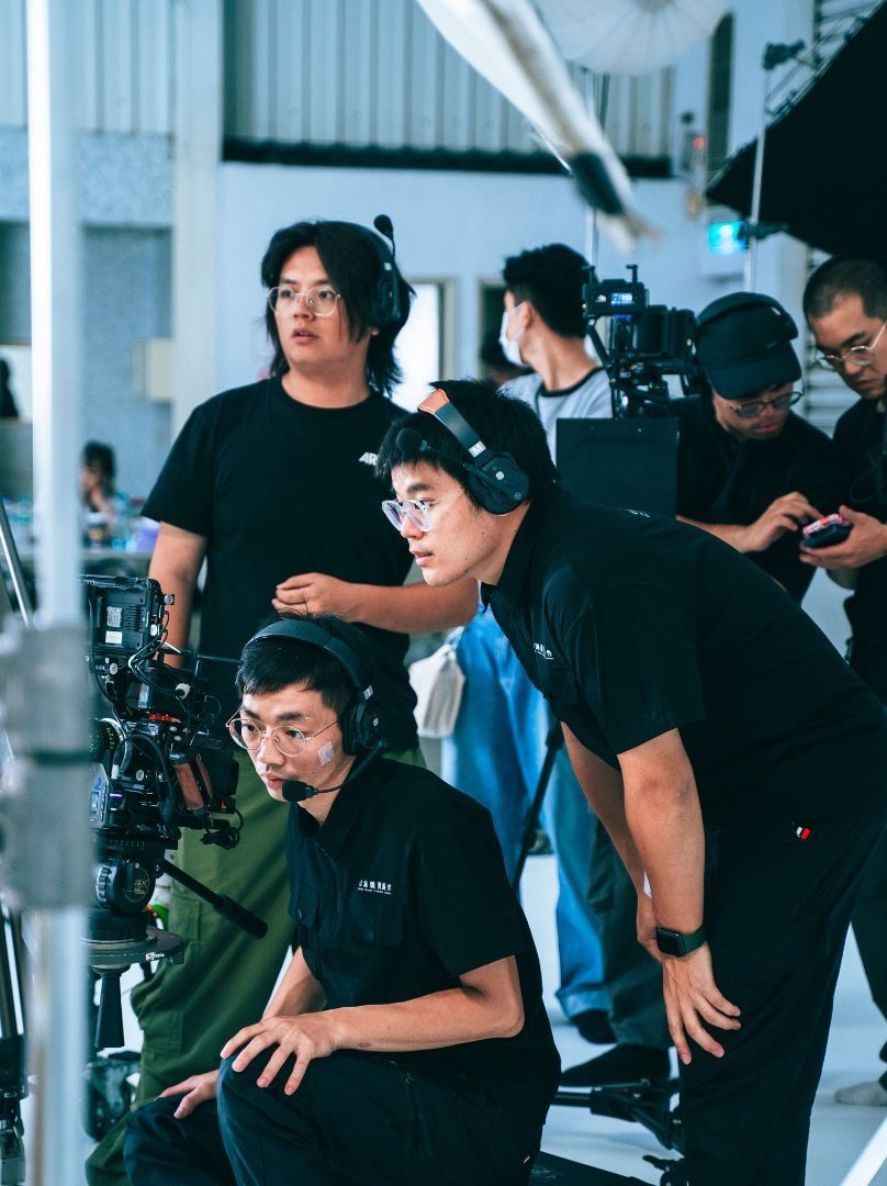Film crew members in black attire operate cameras on a professional video production set indoors.