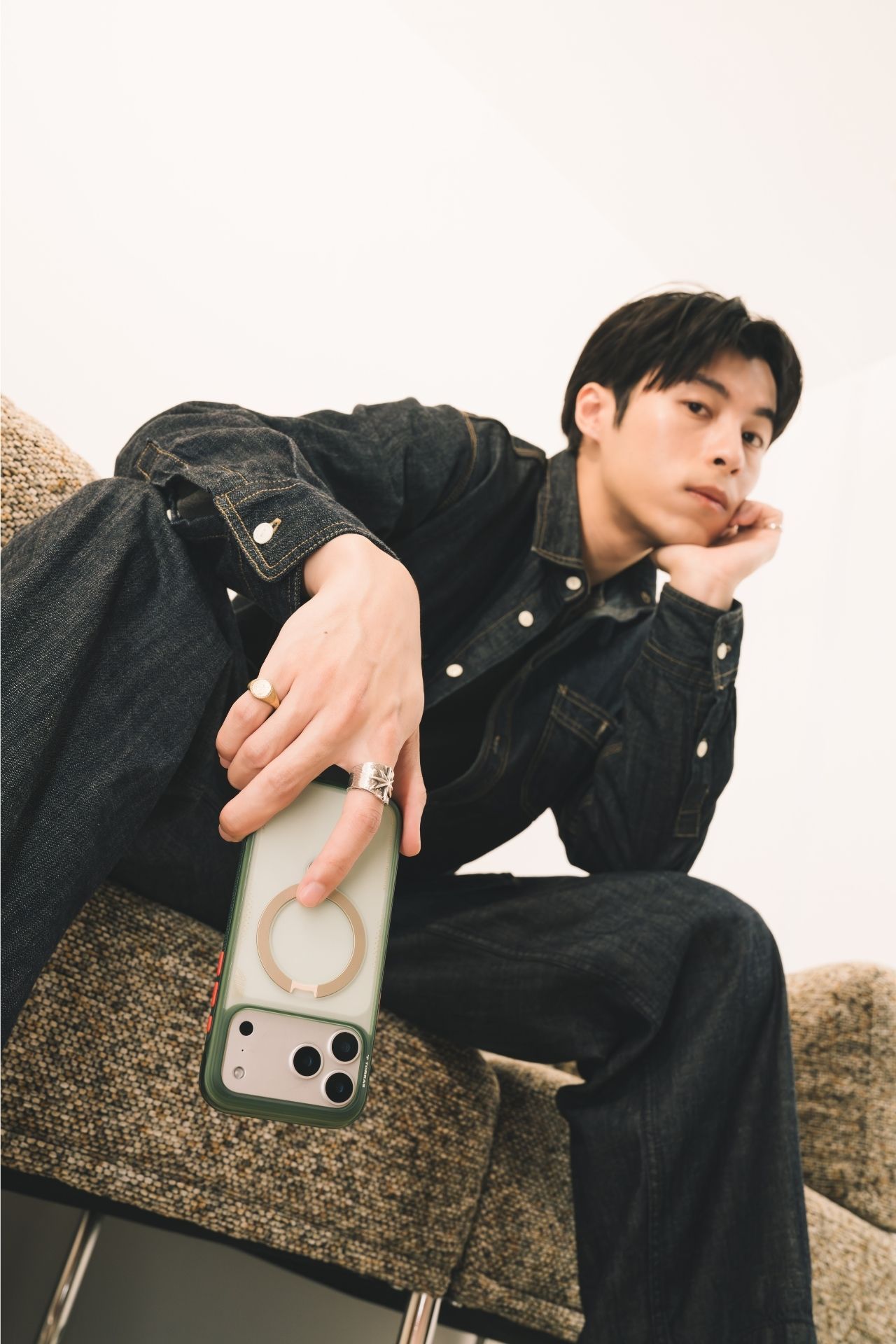 Young man in dark denim outfit sitting casually, holding a green iPhone case with MagSafe ring, wearing gold rings.