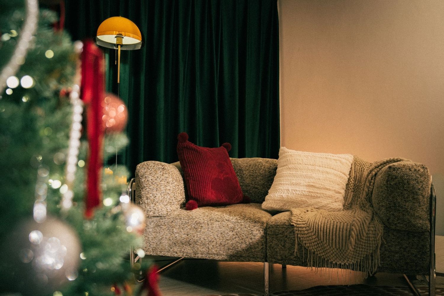 Cozy living room with a gray sofa, red pillows, green curtains, floor lamp, and a decorated Christmas tree in the foreground.