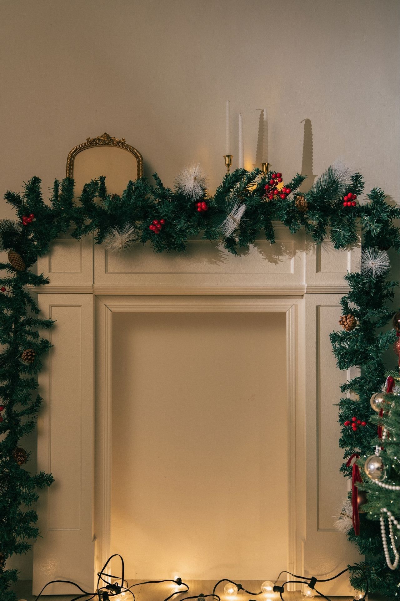 Festive fireplace mantel decorated with Christmas garland, red berries, candles, and warm string lights below.