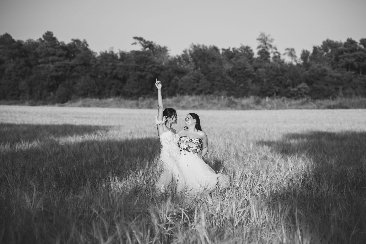 Black and white photo of two brides in a field, one raising her arm joyfully, with trees in the background.