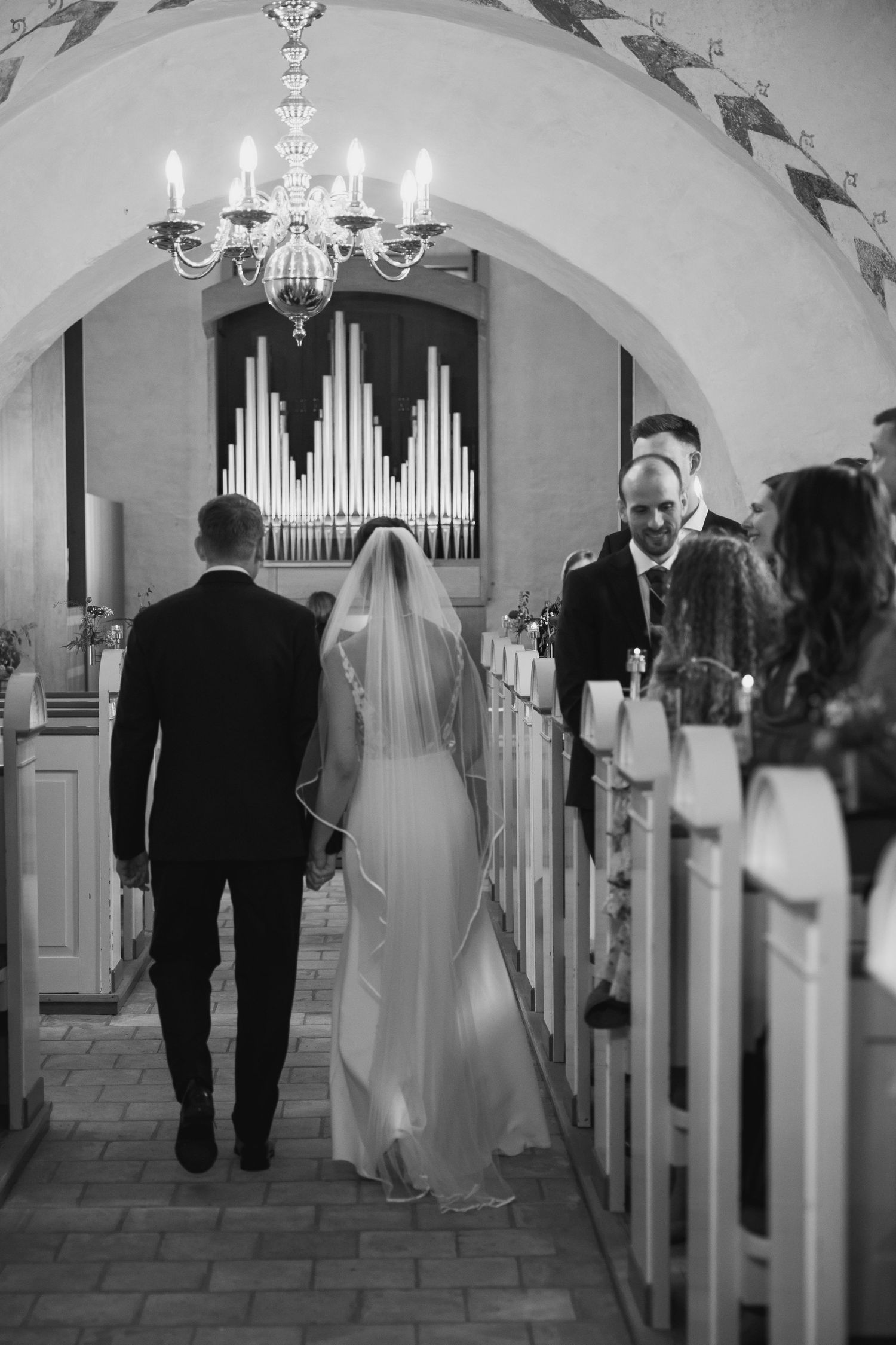 Black and white photo of bride and groom walking down church aisle toward pipe organ during wedding ceremony.