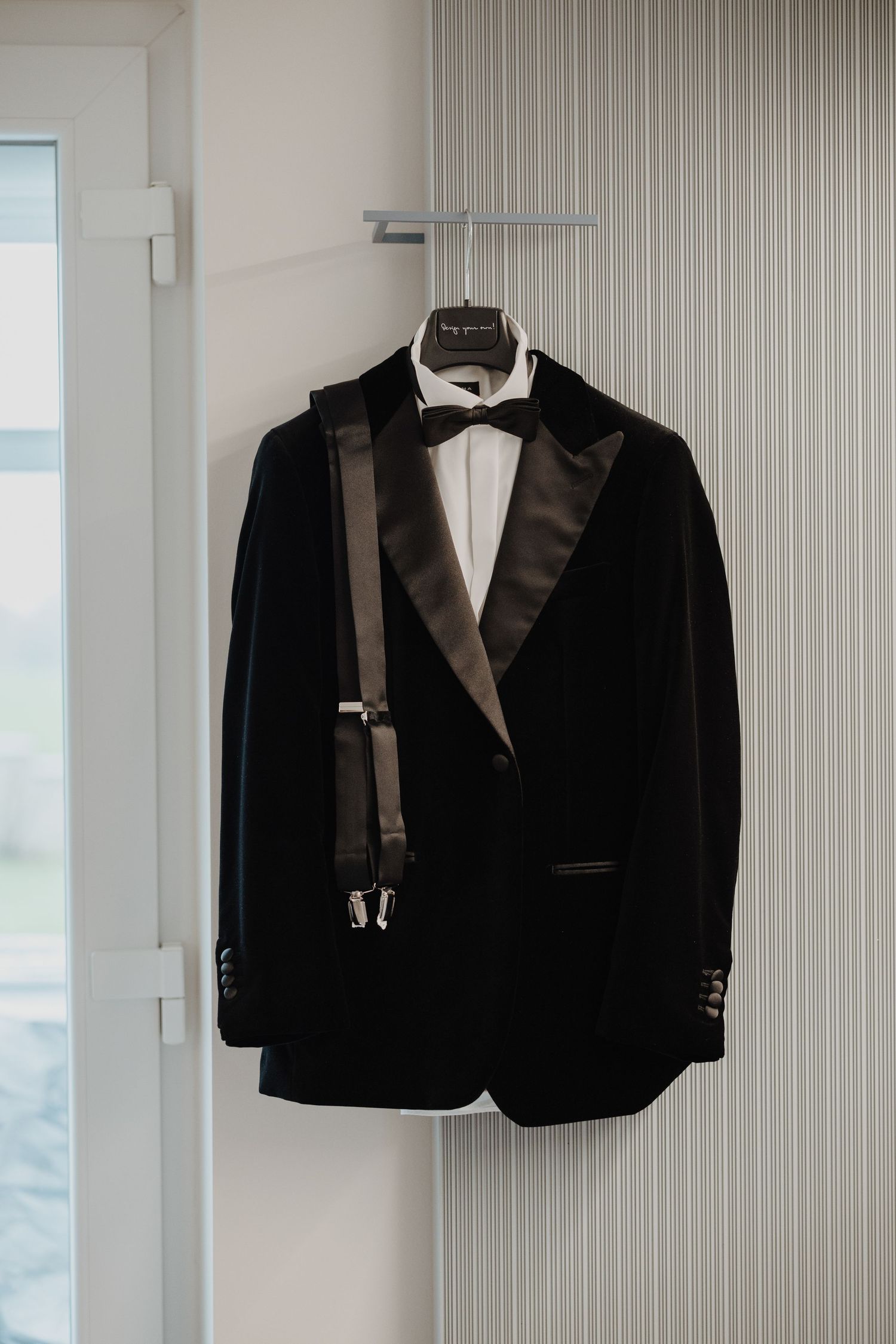 Black velvet tuxedo jacket with satin lapels and suspenders hanging on a door in a bright room.