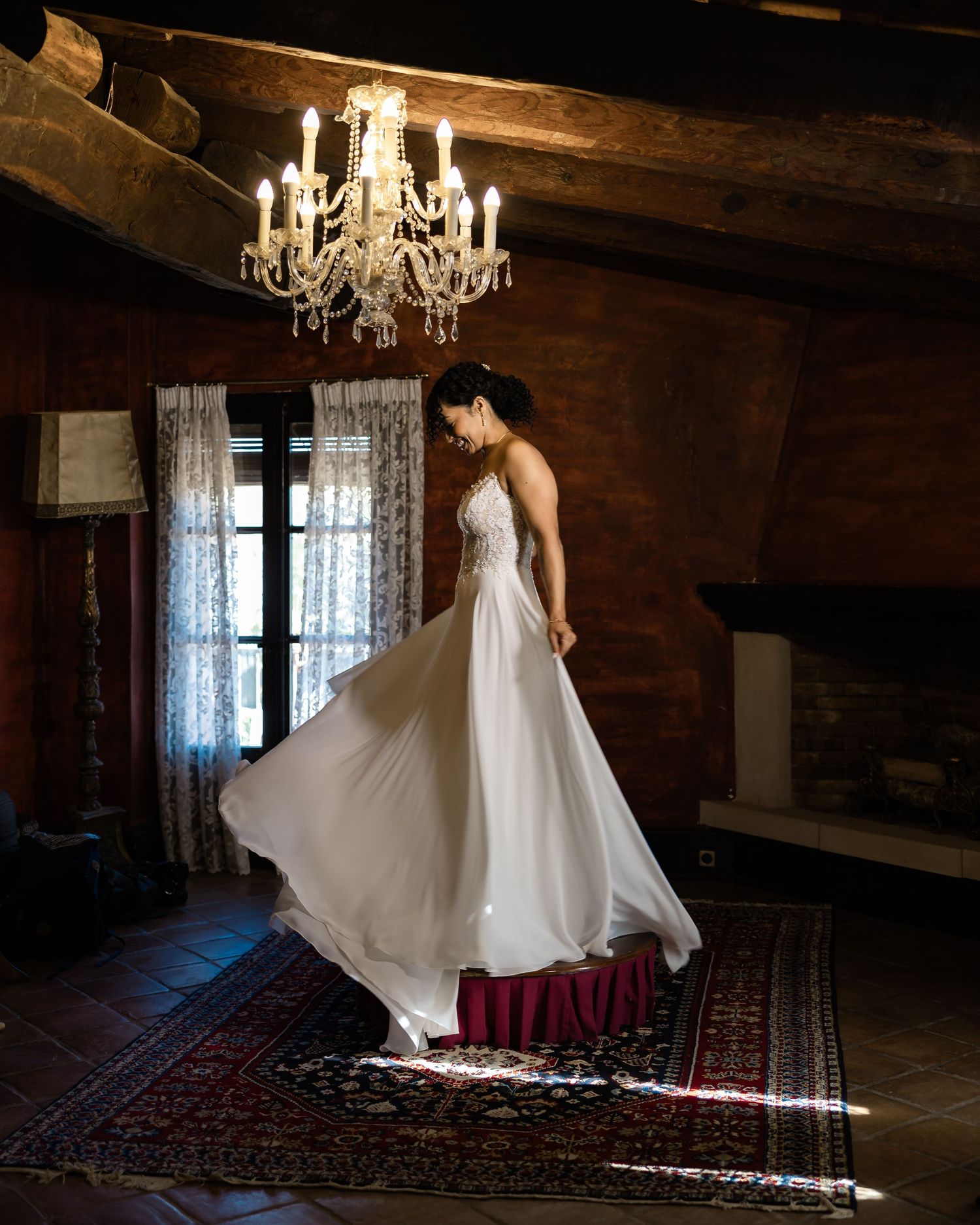 Bride twirling in white gown under crystal chandelier in rustic candlelit room.