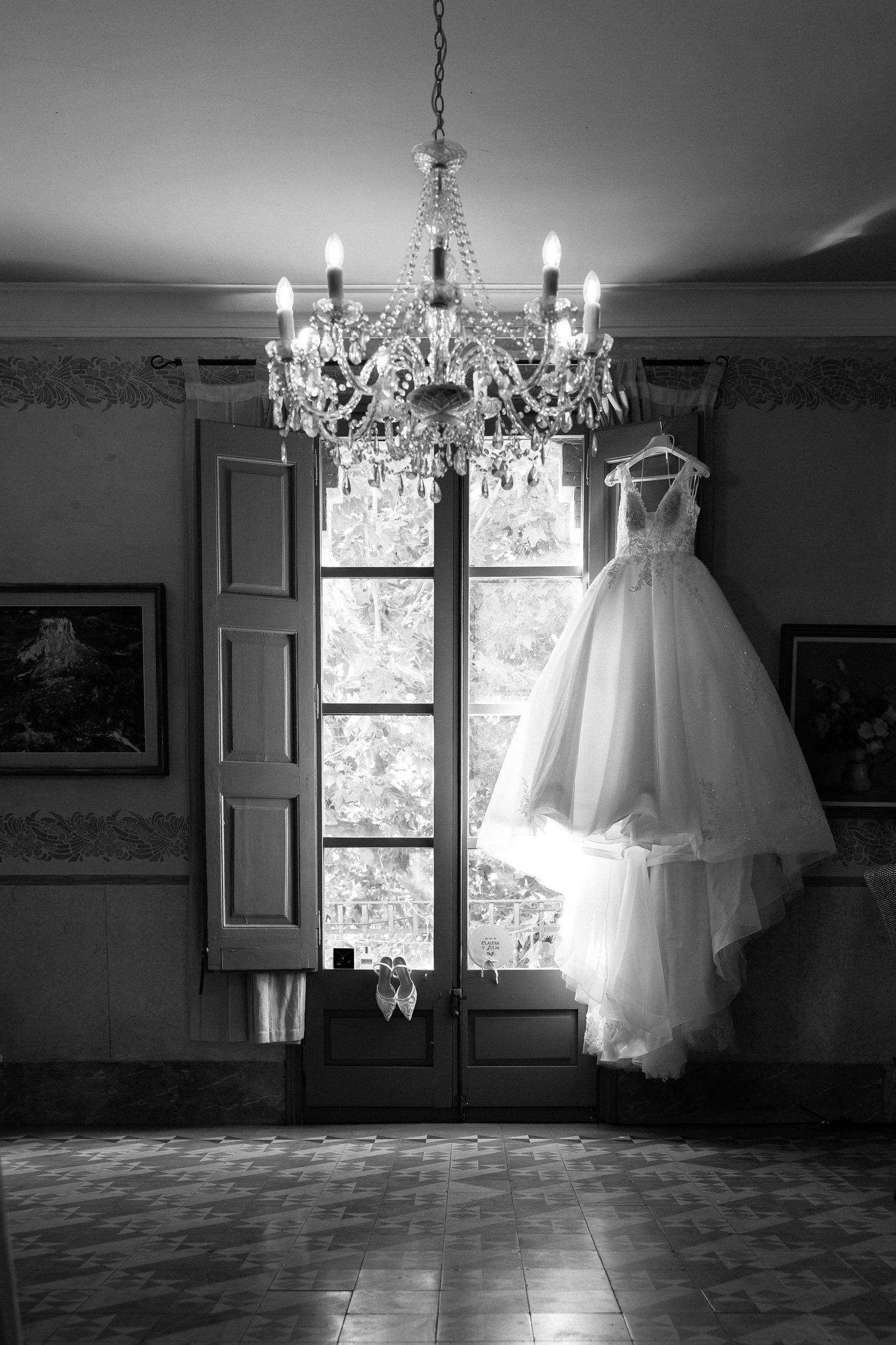 A wedding gown hangs from a crystal chandelier by a tall window in a classic room, black and white.