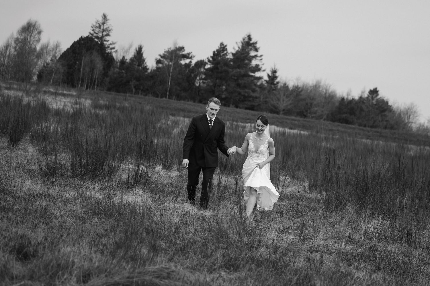 Black and white photo of bride and groom walking hand in hand through a grassy field.