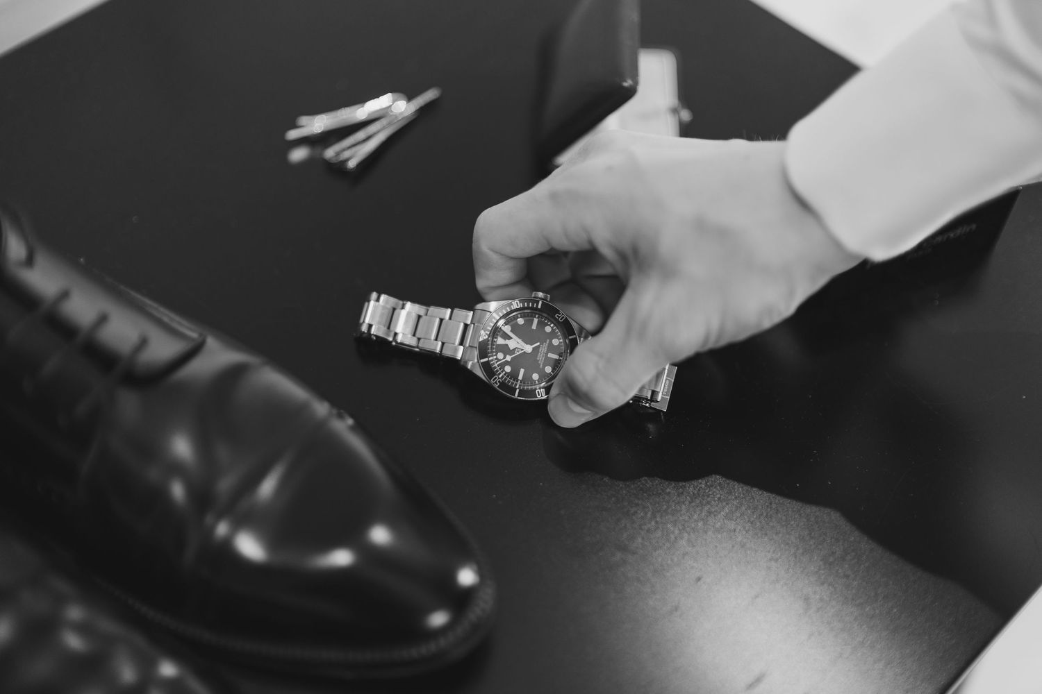 Man holding a silver metal watch beside polished dress shoes on a dark surface in black and white.