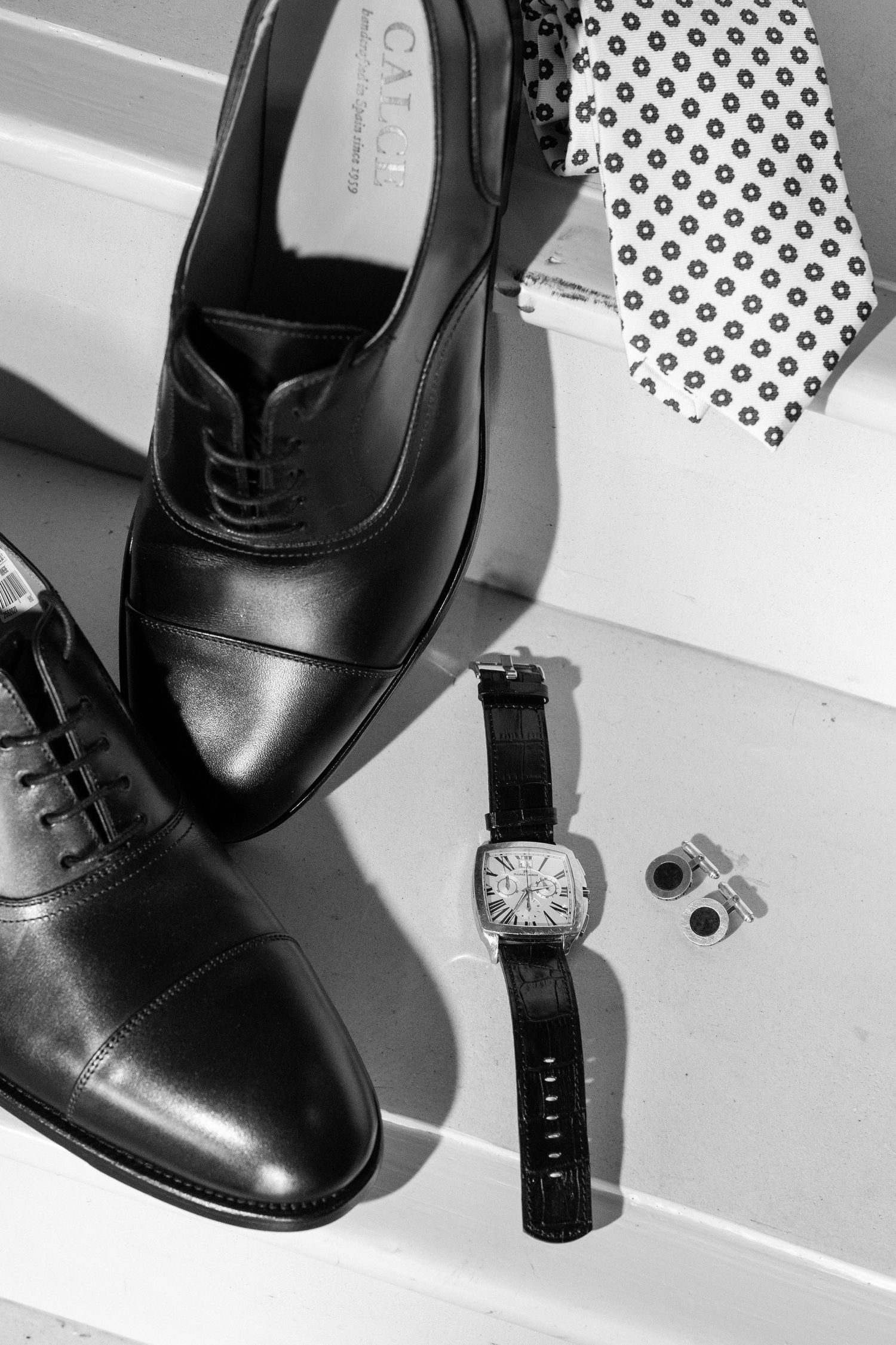 Black leather Oxford shoes with a wristwatch, cufflinks, and patterned tie in a black and white flat lay.