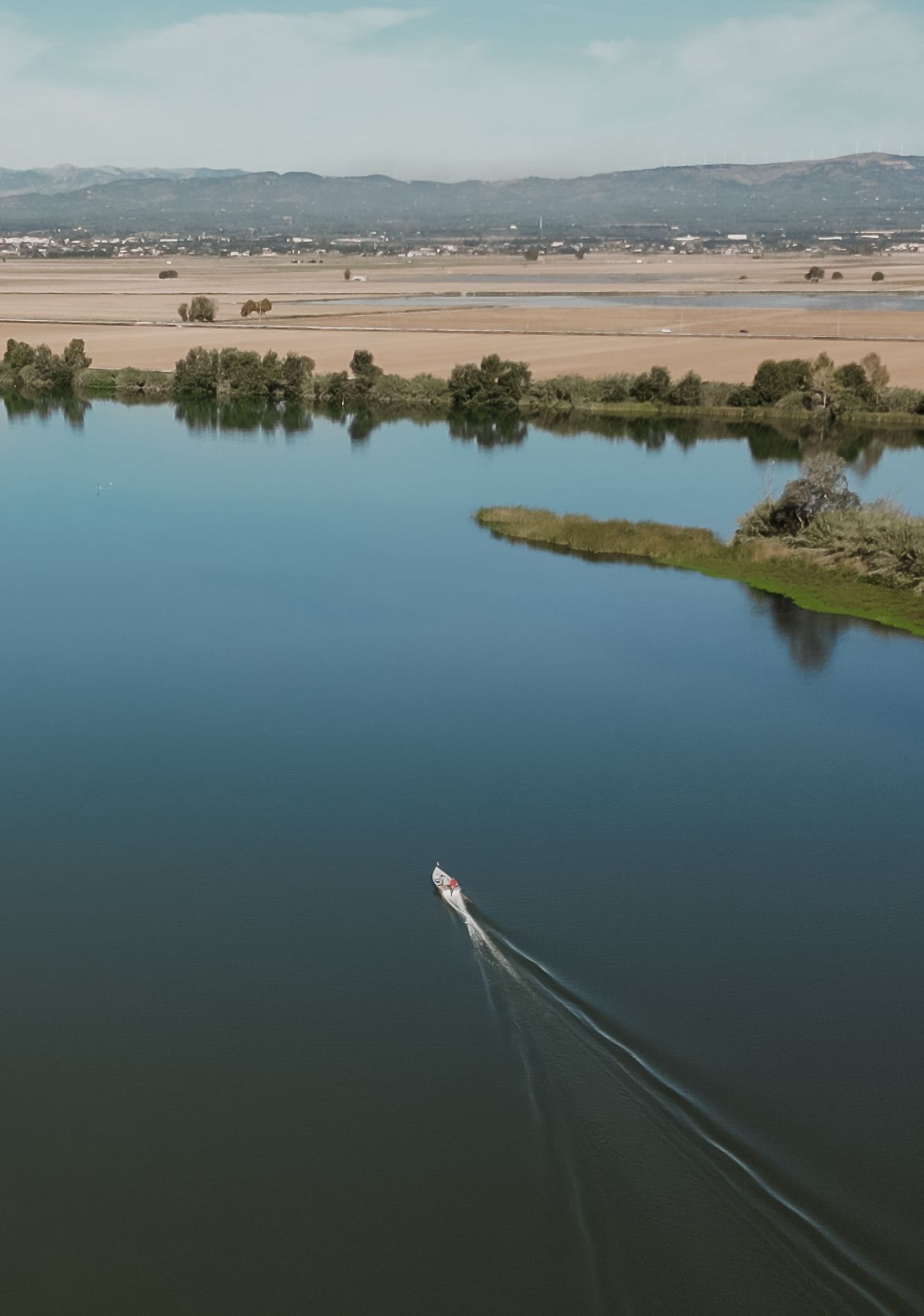 Aerial drone view of a small boat leaving a wake across a calm river delta landscape.