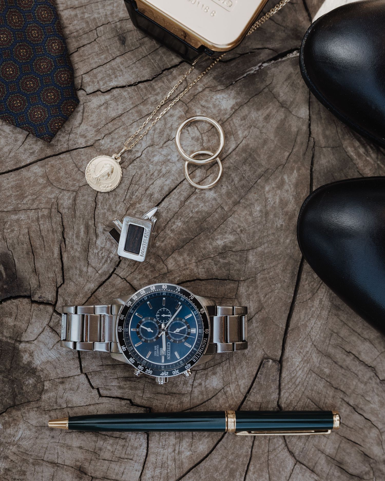 Flat lay of groom wedding accessories including watch, rings, tie, pen, cufflinks and dress shoes.