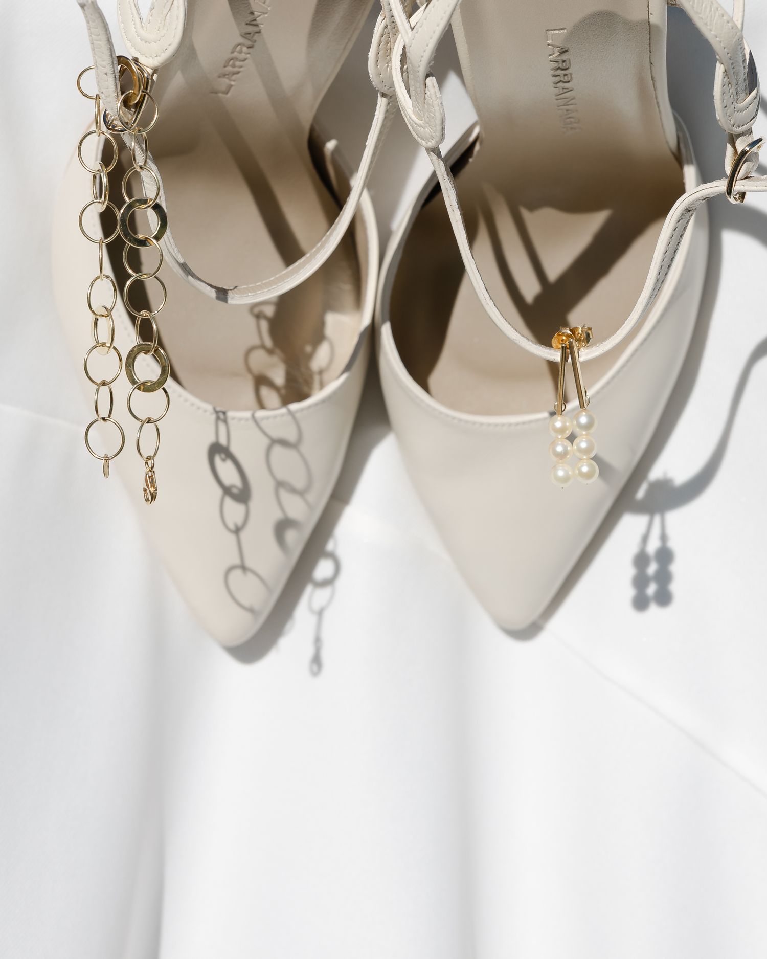 Elegant white bridal heels with gold chain and pearl shoe clips on white fabric background.