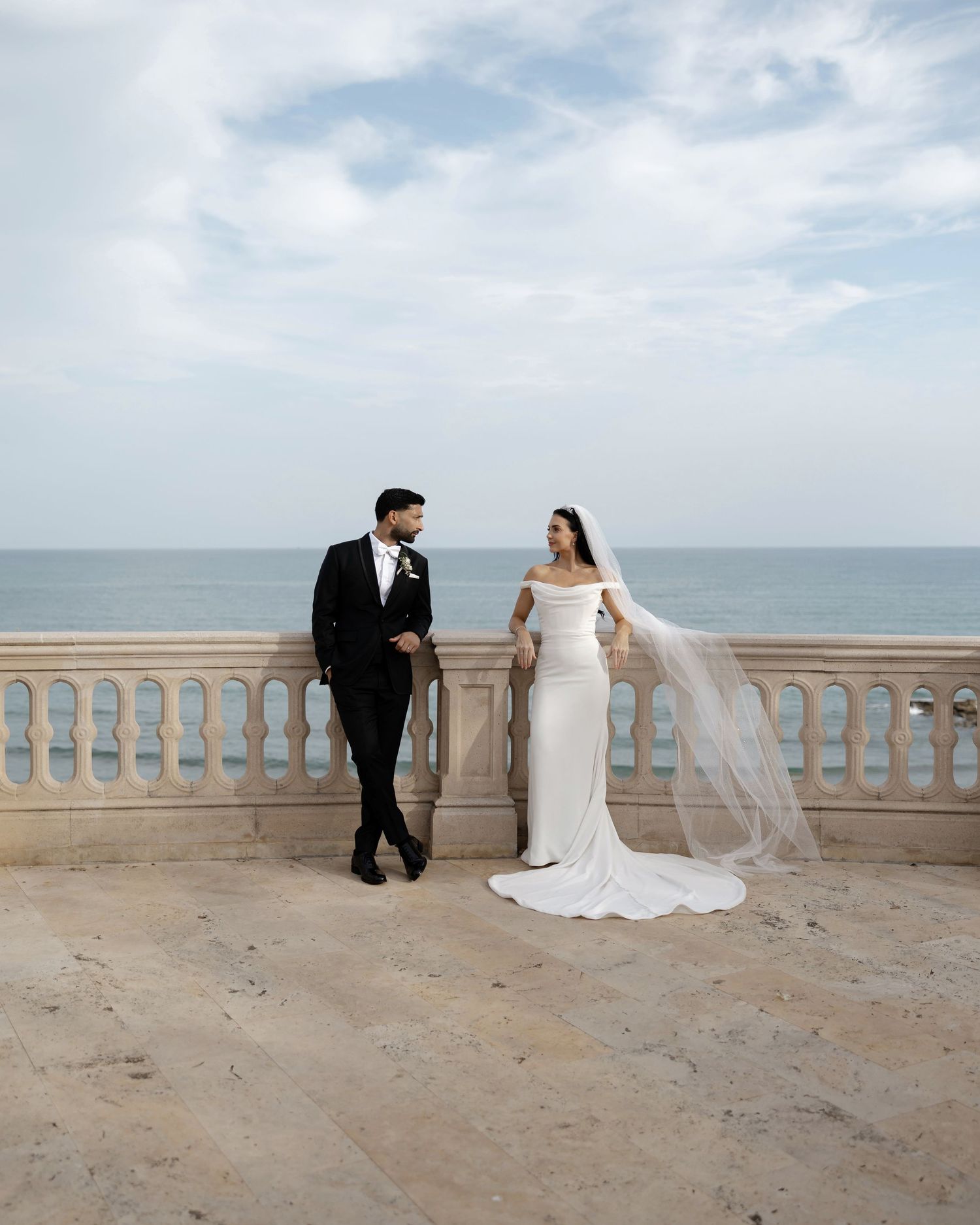 Bride and groom stand on oceanfront terrace with white balustrade and sea view.