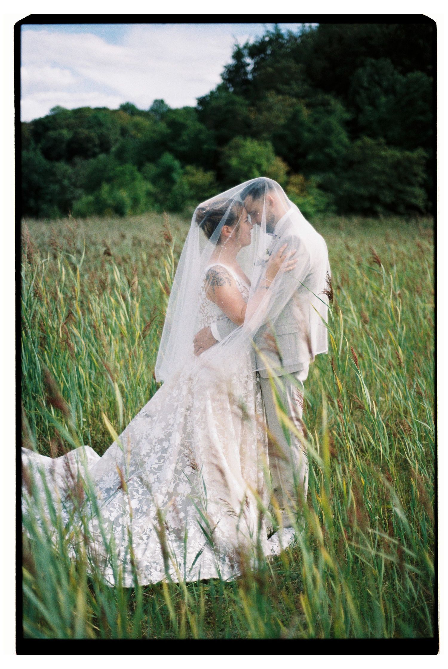 Bride and groom kissing under a veil in a lush green field, wearing a lace wedding gown with a long train.