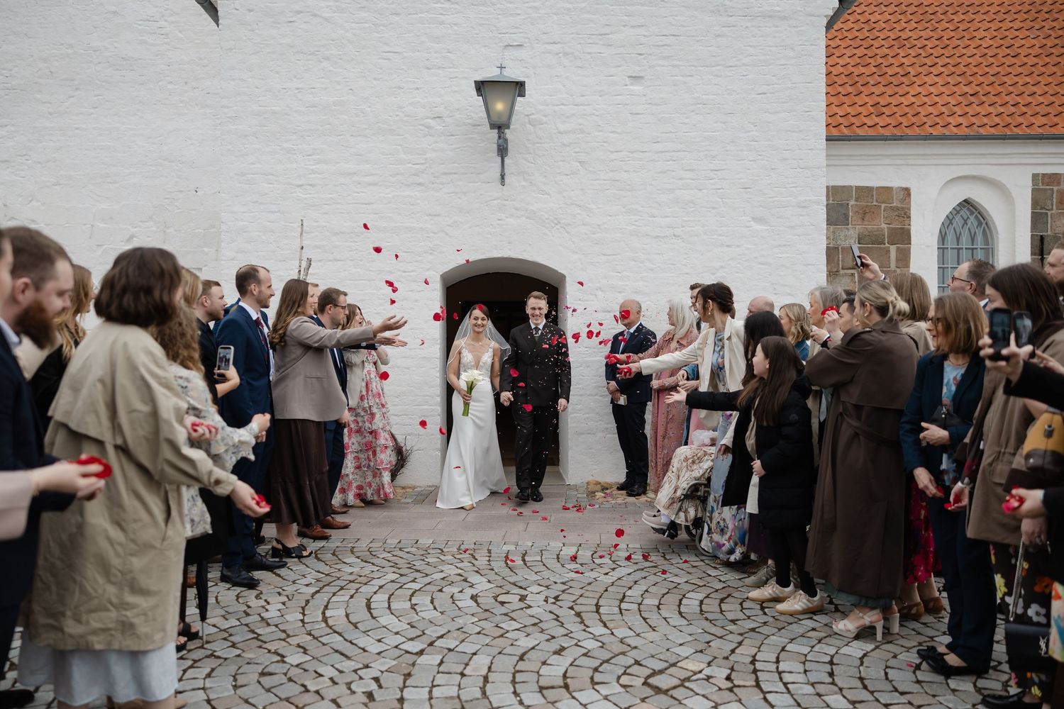 Bride and groom exit white stone church as guests throw red rose petals on cobblestone pathway.