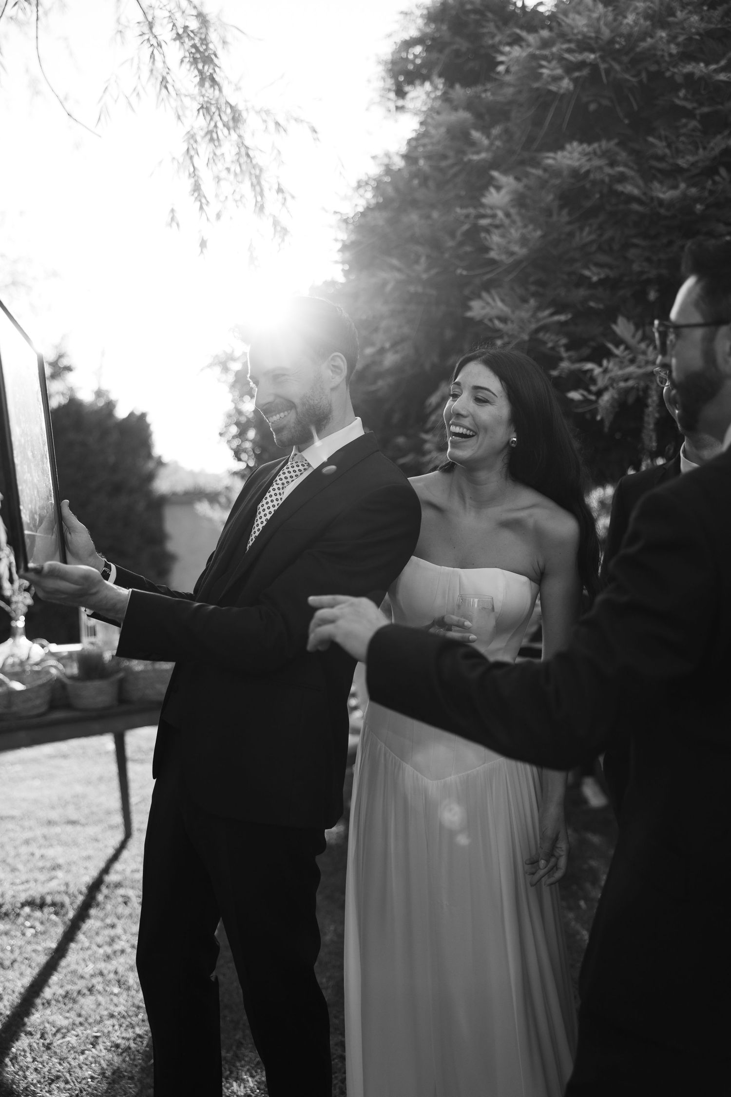 Joyful bride in white gown laughing with groom at outdoor wedding reception with sunburst effect.