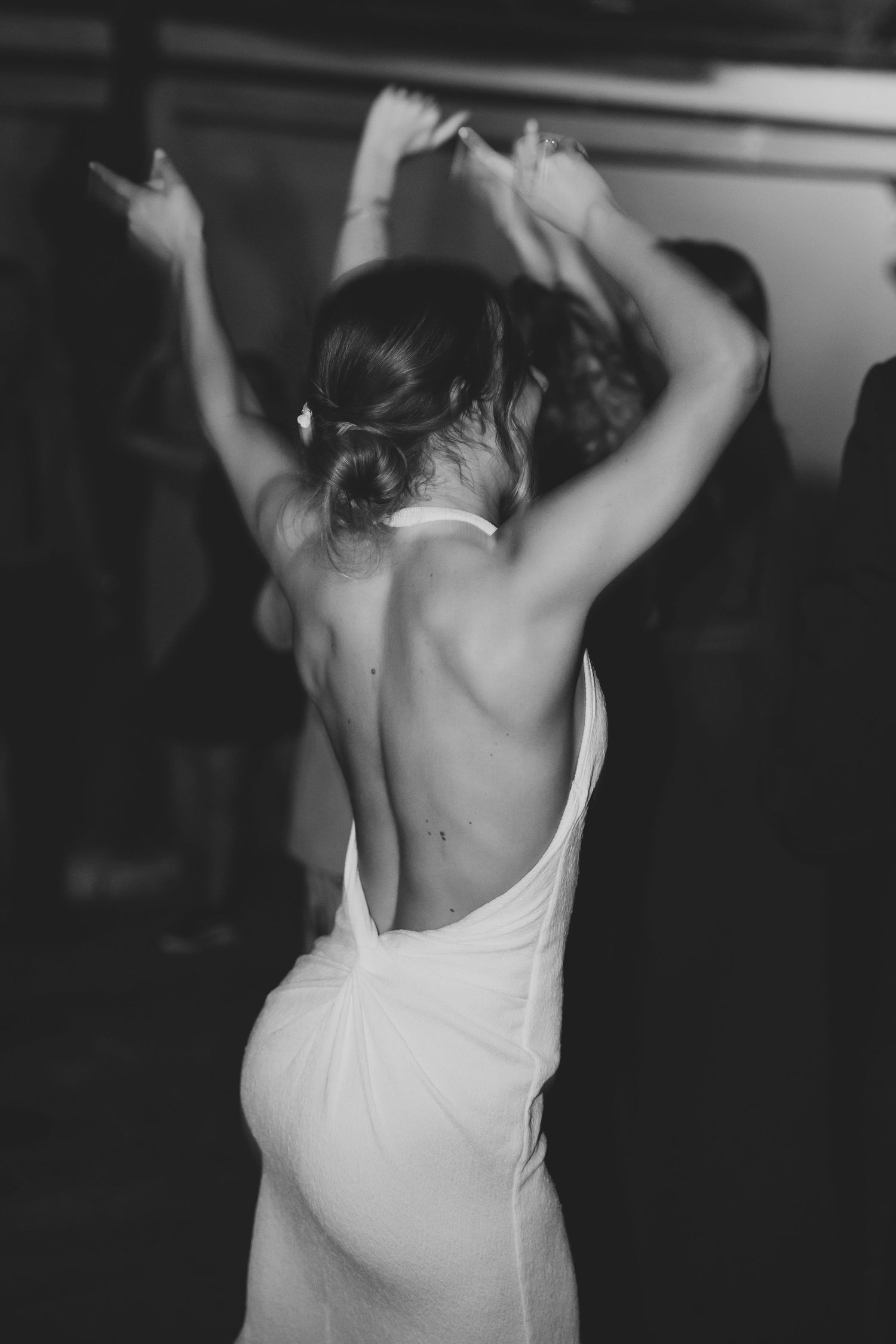 Black and white photo of bride in backless halter gown dancing with arms raised at wedding reception.