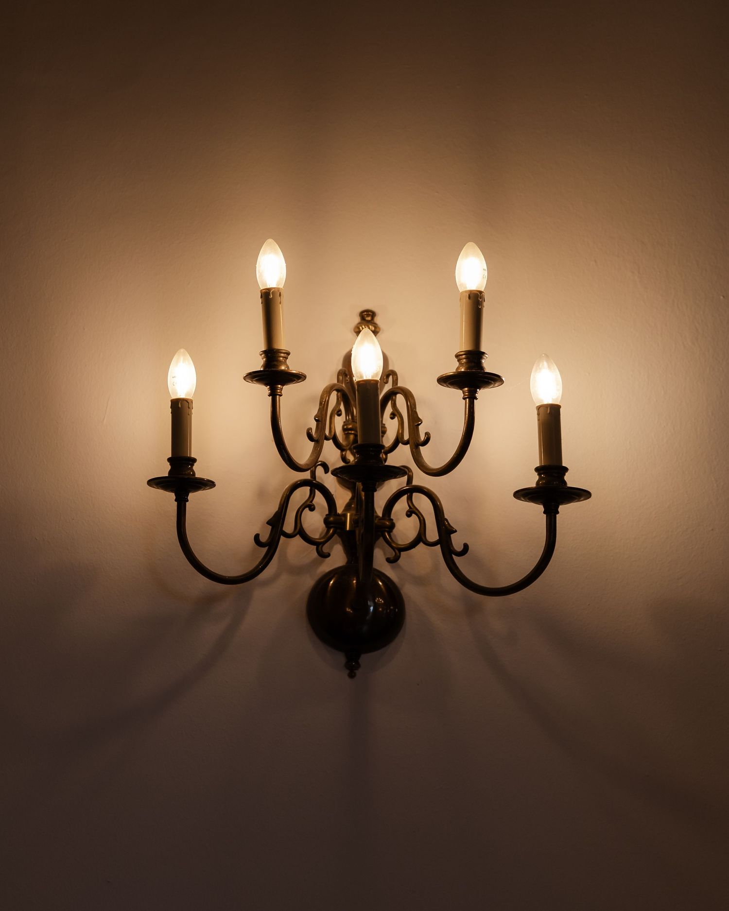Elegant antique brass five-arm candelabra wall sconce with lit candle bulbs on warm wall.
