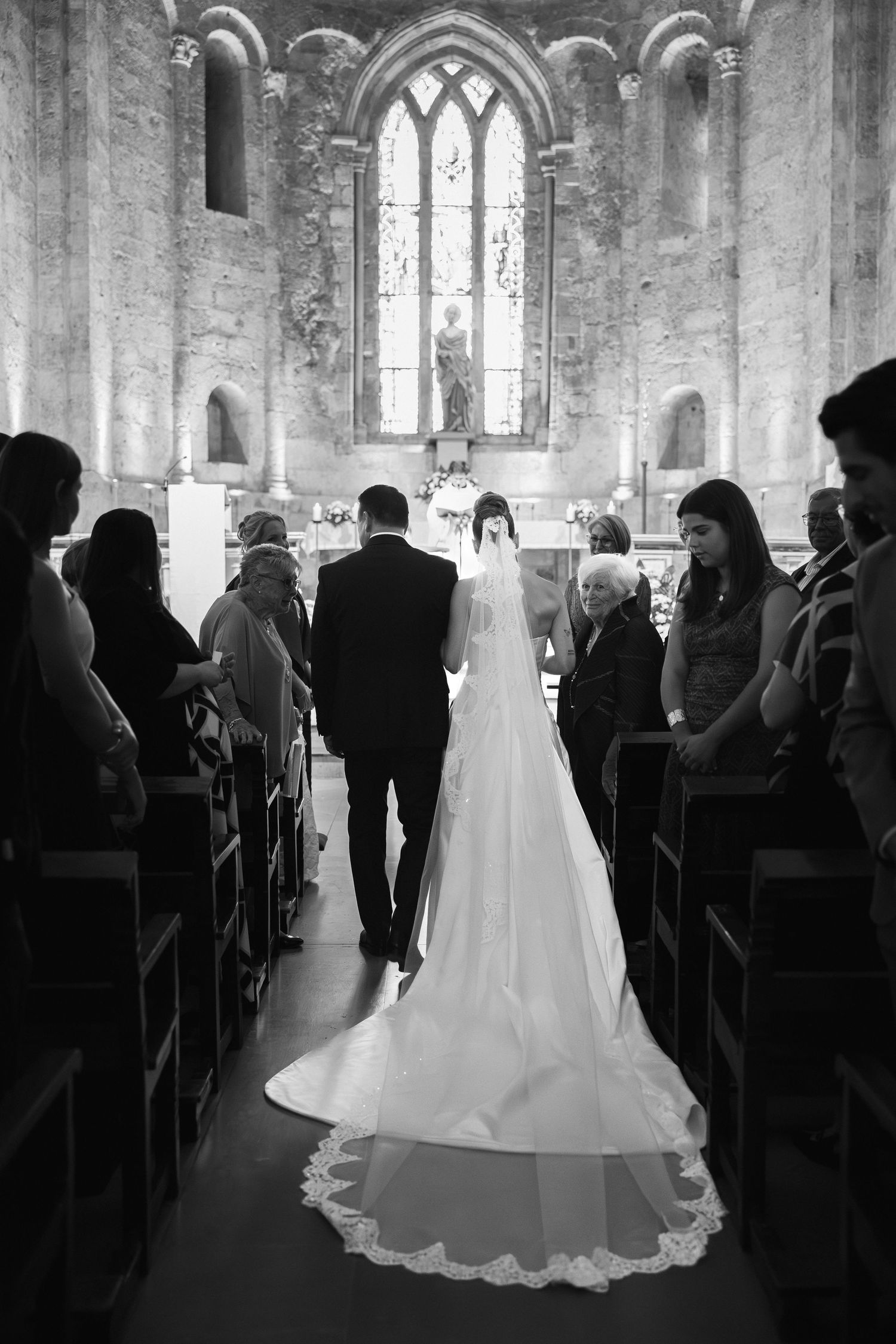 Black and white photo of bride and groom walking down church aisle with guests seated in historic stone chapel.
