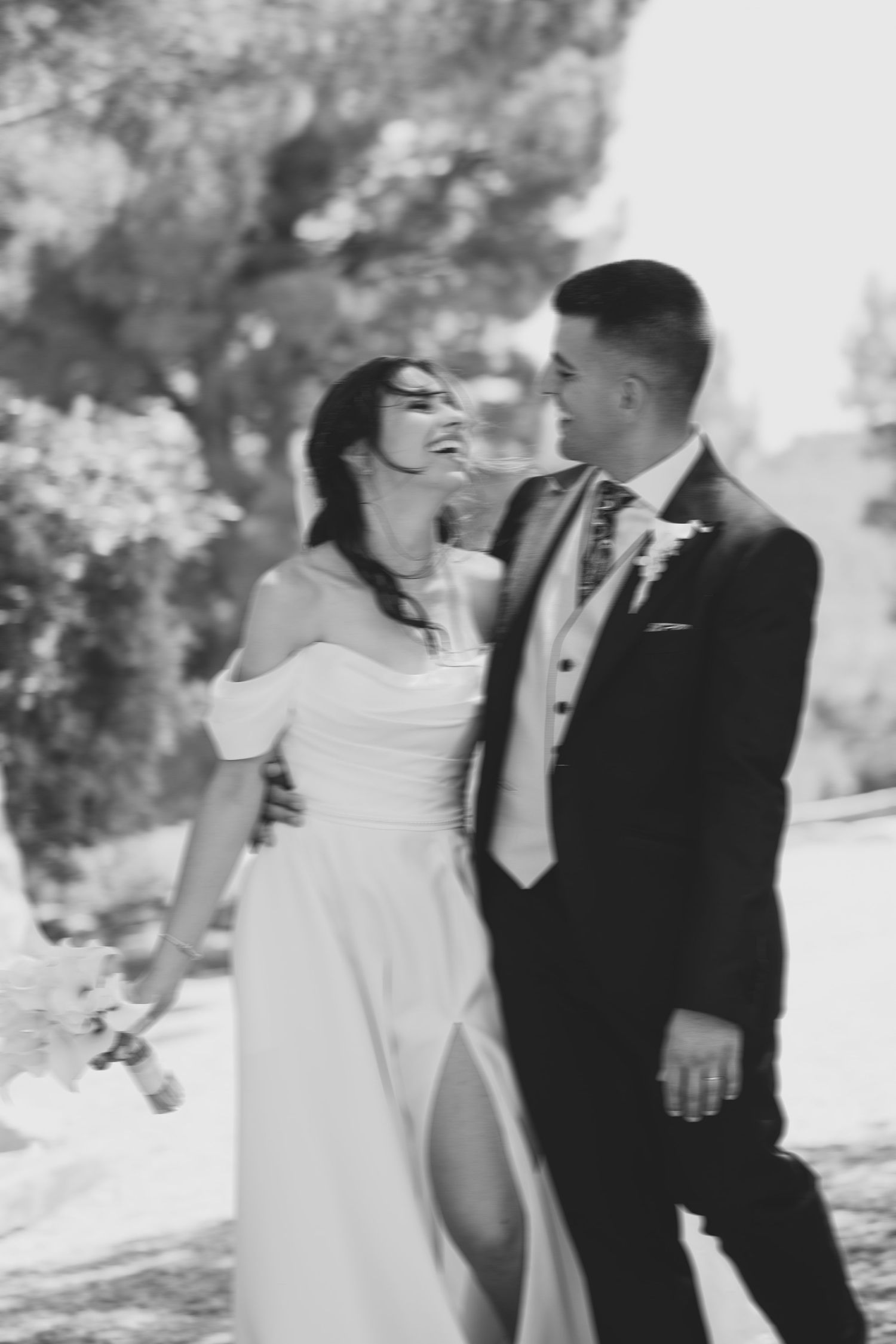 Black and white photo of laughing bride in off-shoulder gown walking with groom in suit outdoors.