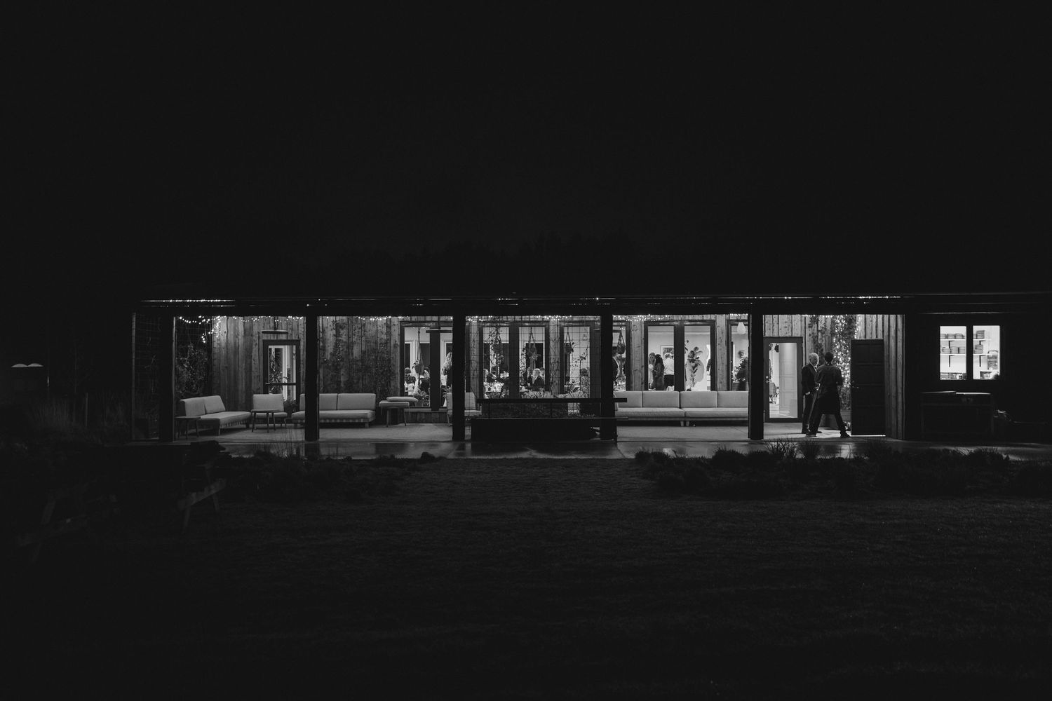 Black and white nighttime exterior of illuminated modern building with silhouetted figure outside.