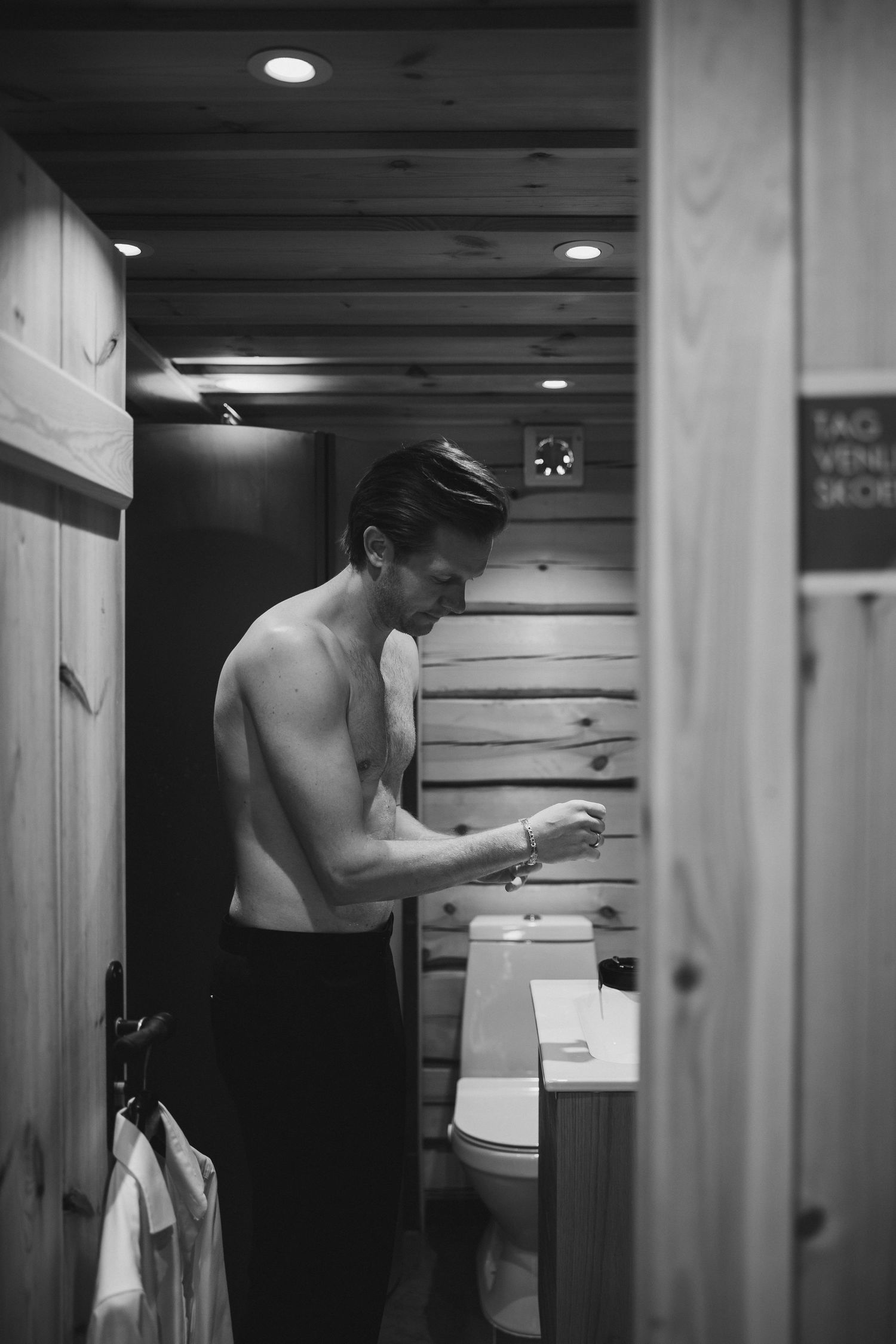 Black and white photo of shirtless man washing hands in a wooden cabin bathroom with recessed lighting.