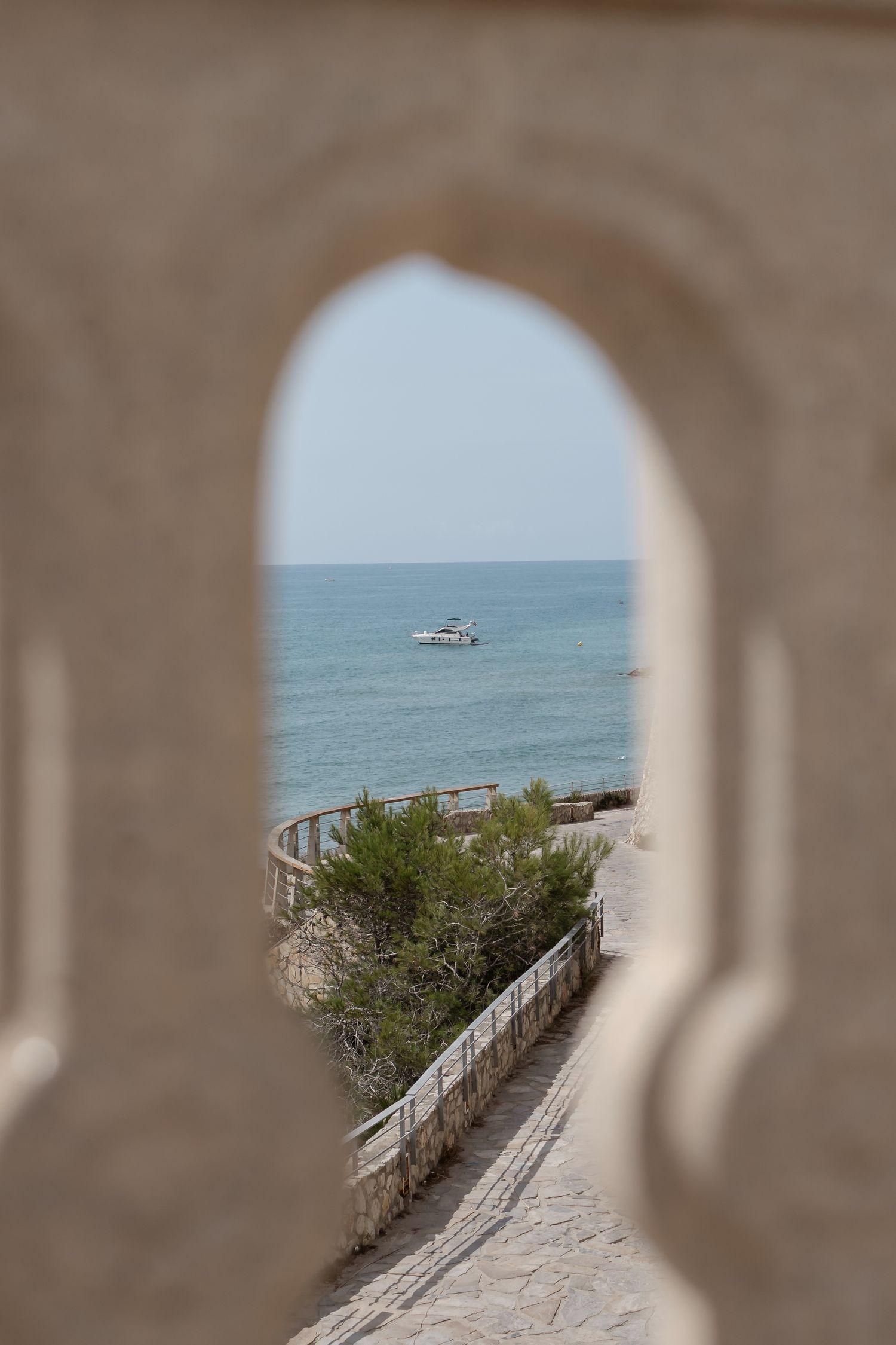 Moorish arch frames a scenic coastal pathway leading to a calm blue sea with a white boat in the distance.
