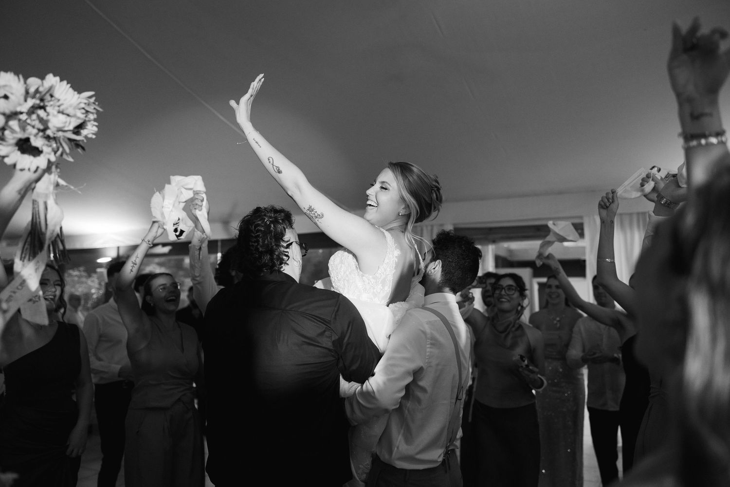 Bride being lifted by guests during wedding reception celebration in black and white photography.