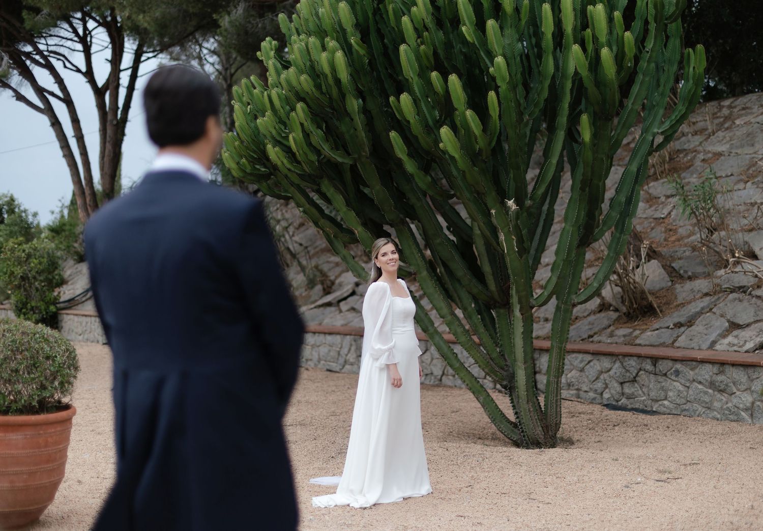 Bride in white gown stands before a tall cactus as groom watches in a garden setting.