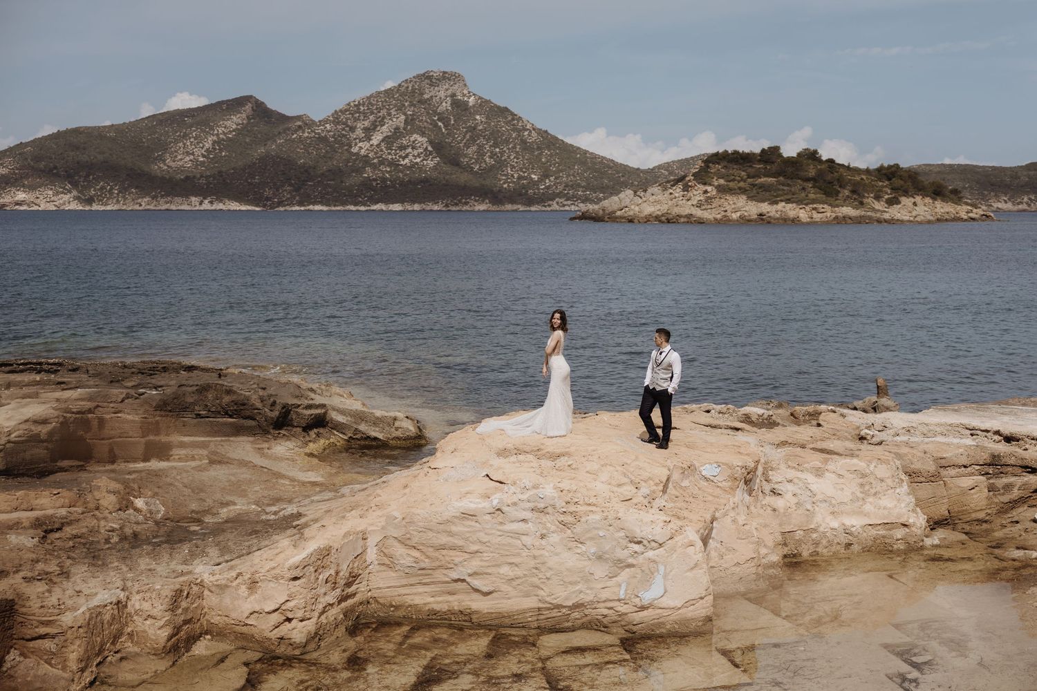 Bride and groom standing on rocky coastal landscape with mountains and blue water behind them.