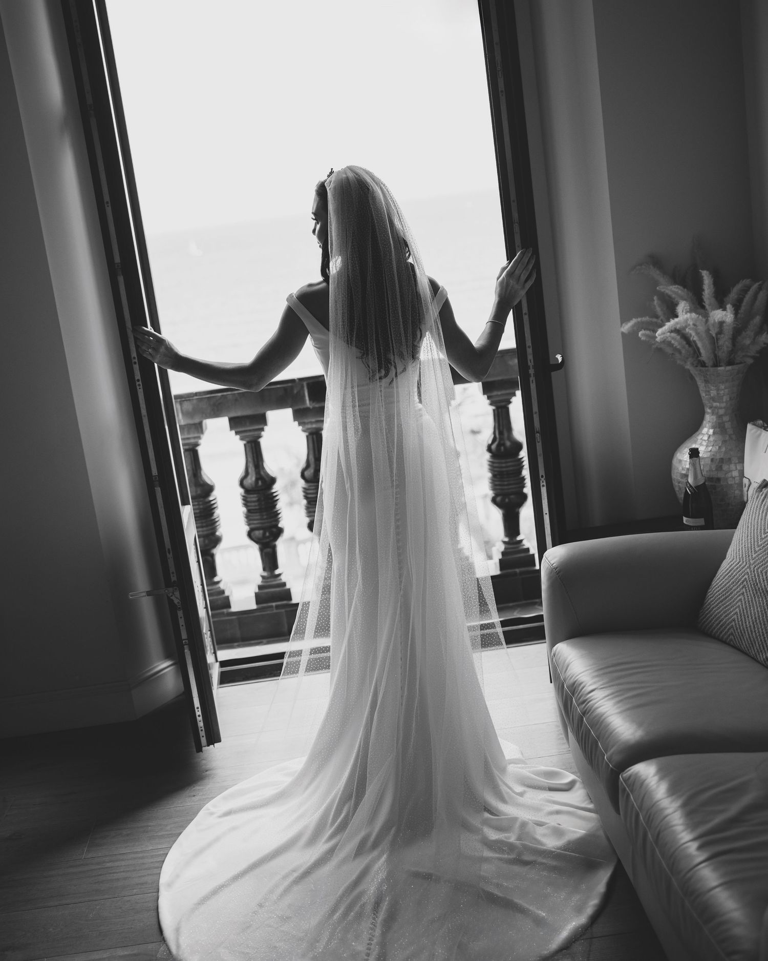 Black and white silhouette of a bride in a flowing veil standing at an open balcony doorway in a grand room.