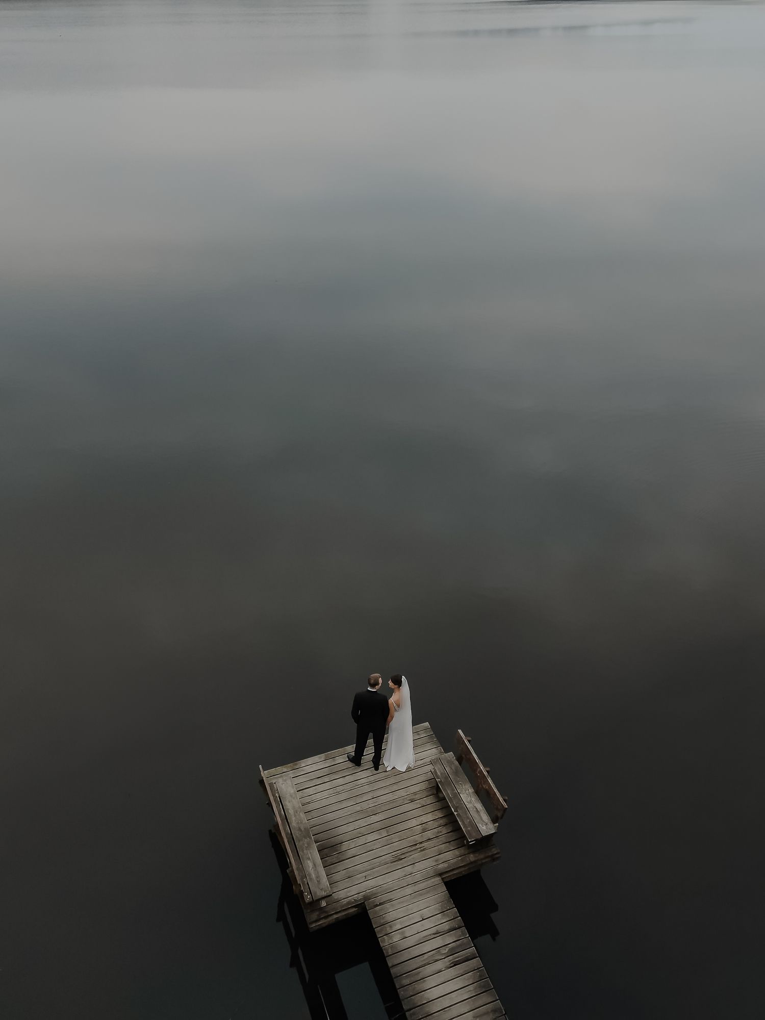 Aerial view of wedding couple standing on wooden dock over calm misty gray lake.