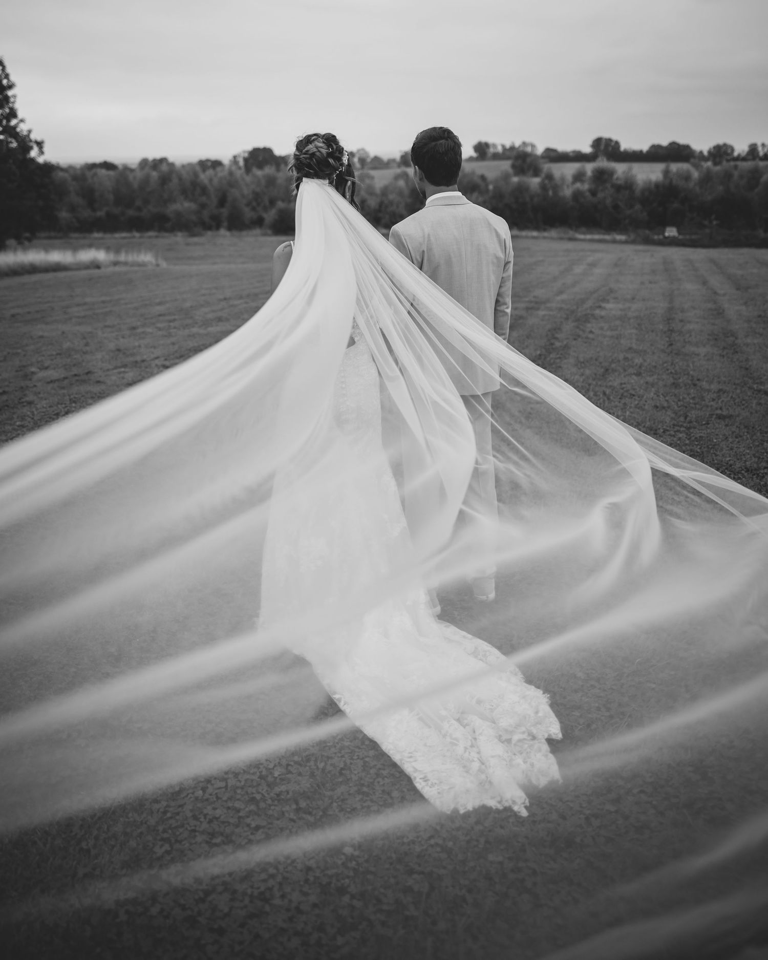 Black and white photo of bride and groom walking in field with dramatic flowing cathedral wedding veil.