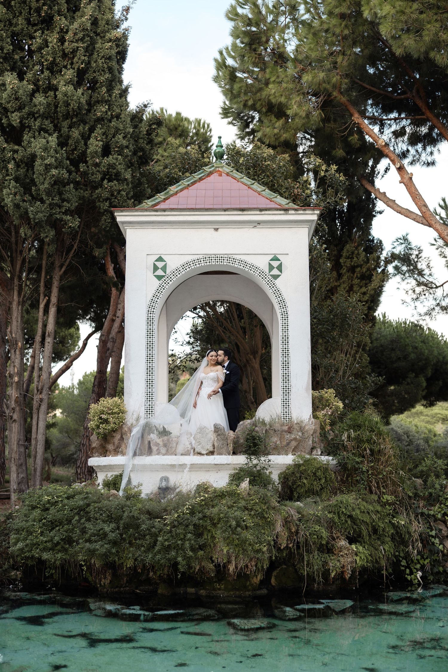 Bride and groom pose inside a white ornate garden pavilion surrounded by lush trees and a tranquil pond.